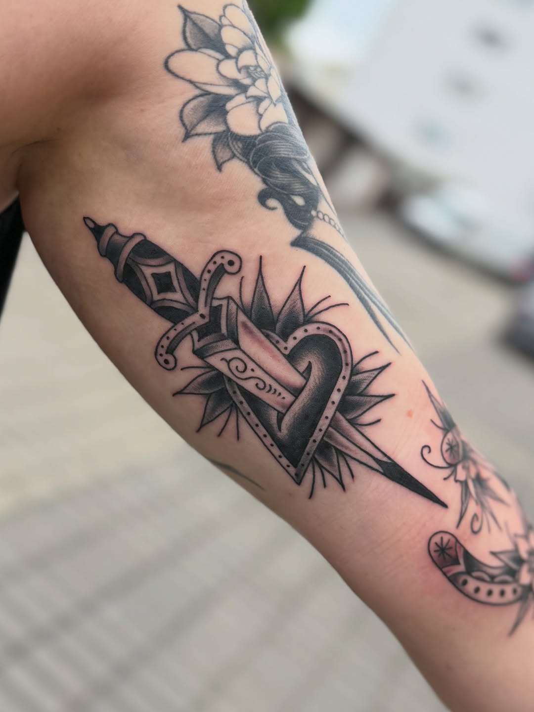 Dagger piercing a heart with radiating spikes surrounded by ornamental scrollwork, displayed on an arm in black and grey ink.