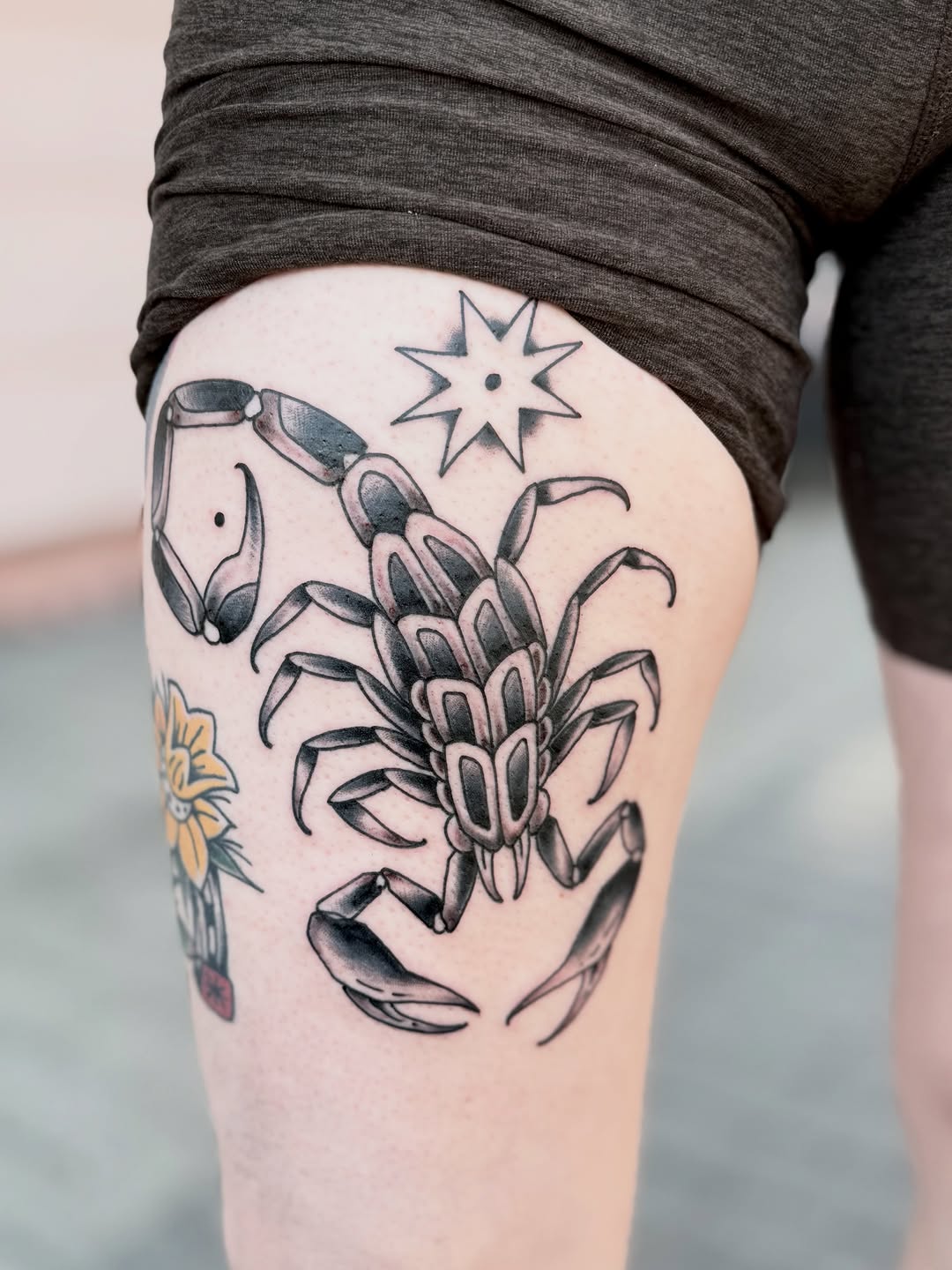 Detailed scorpion tattoo with star and botanical elements in black ink on thigh, shown on pale skin.