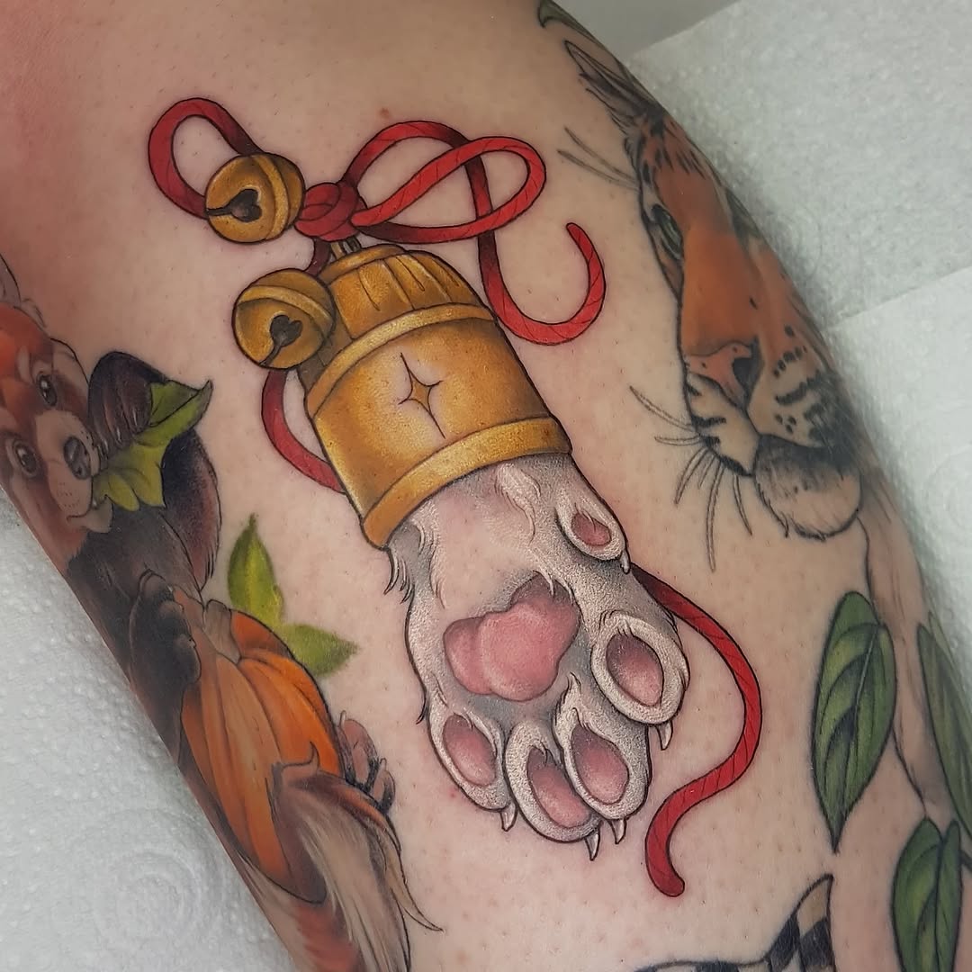 Colorful arm tattoo featuring a paw pad with pink beans, golden bells tied with red ribbon, and surrounding floral elements.