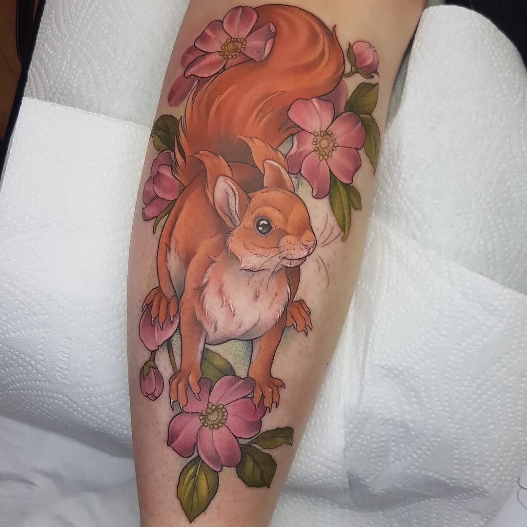 Orange squirrel holding fluffy tail surrounded by pink flowers with green leaves, tattooed on upper leg.