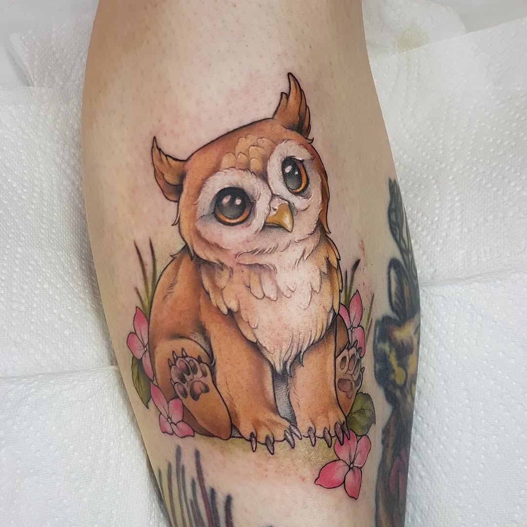 Brown and cream owl with large eyes surrounded by pink flowers and green foliage, tattooed on the calf.