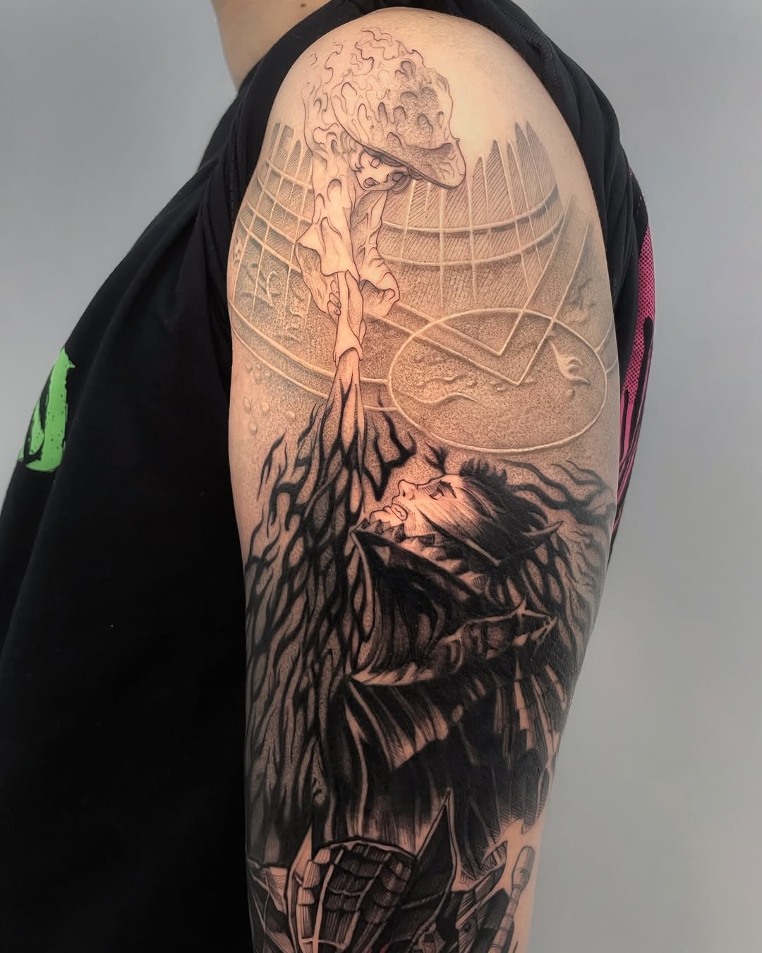 Shoulder tattoo featuring a figure with a large moon head wearing a hat, with roots extending downward and a second face below, rendered in black and gray ink.