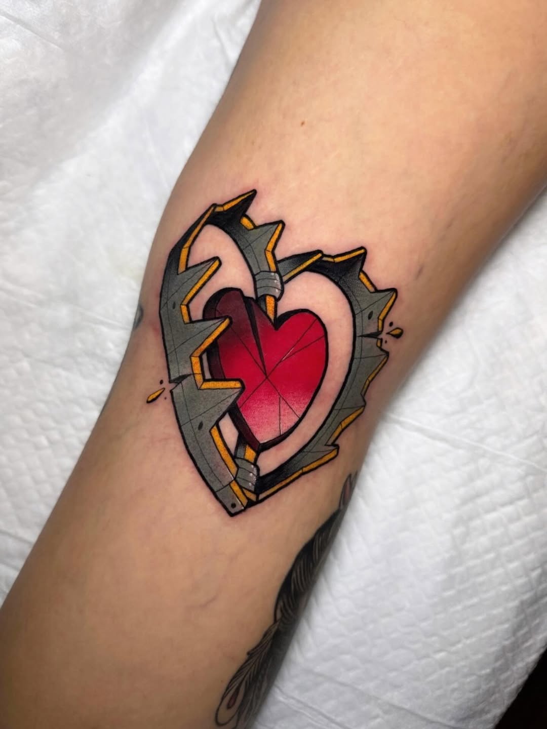 Mechanical armored heart tattoo on forearm with gray metal plating, gold accents, and bright red center heart, featuring black outlines.