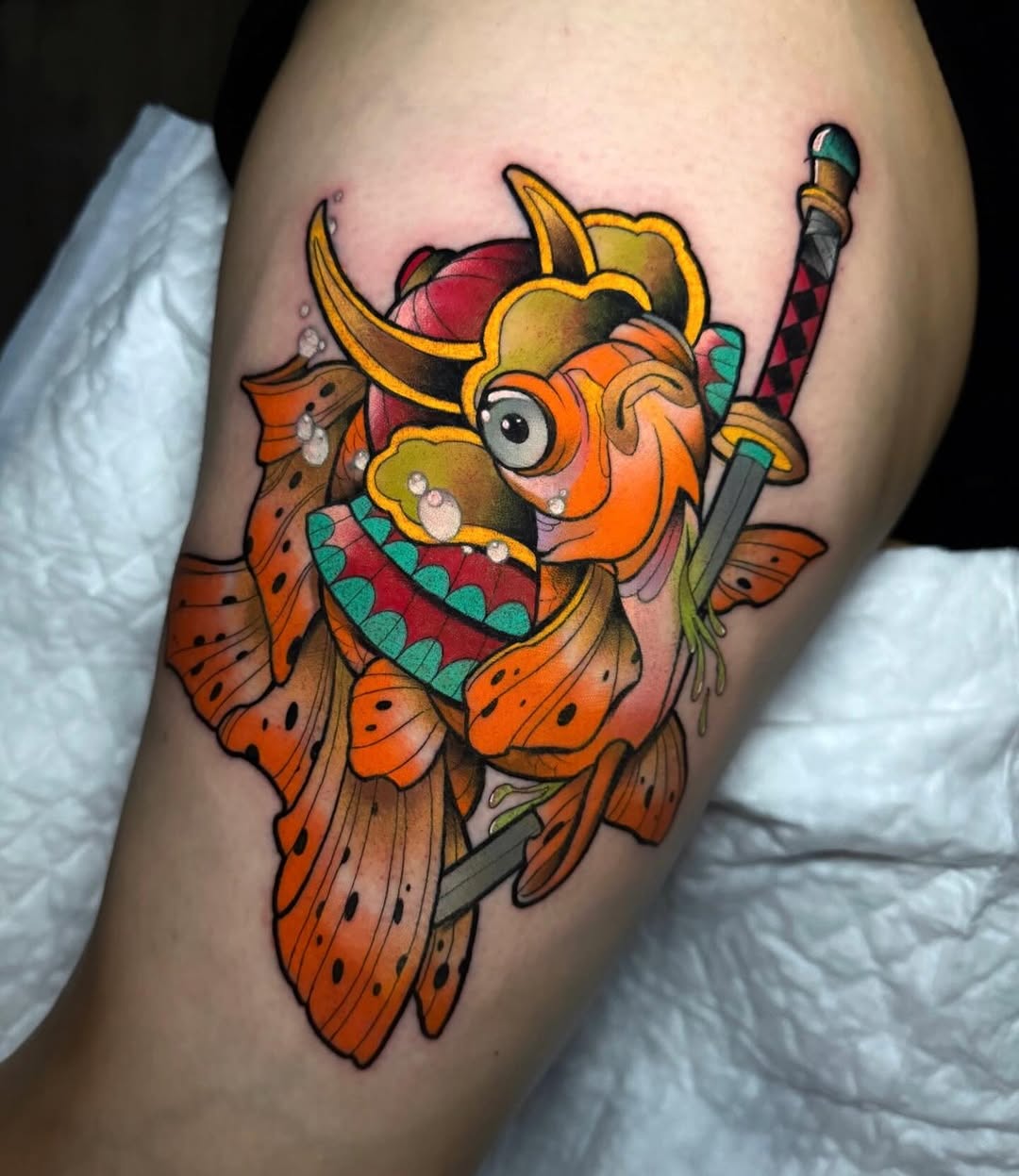 Colorful koi fish tattoo on upper arm featuring orange, yellow, red, and teal scales, wielding swords with detailed shading and line work.