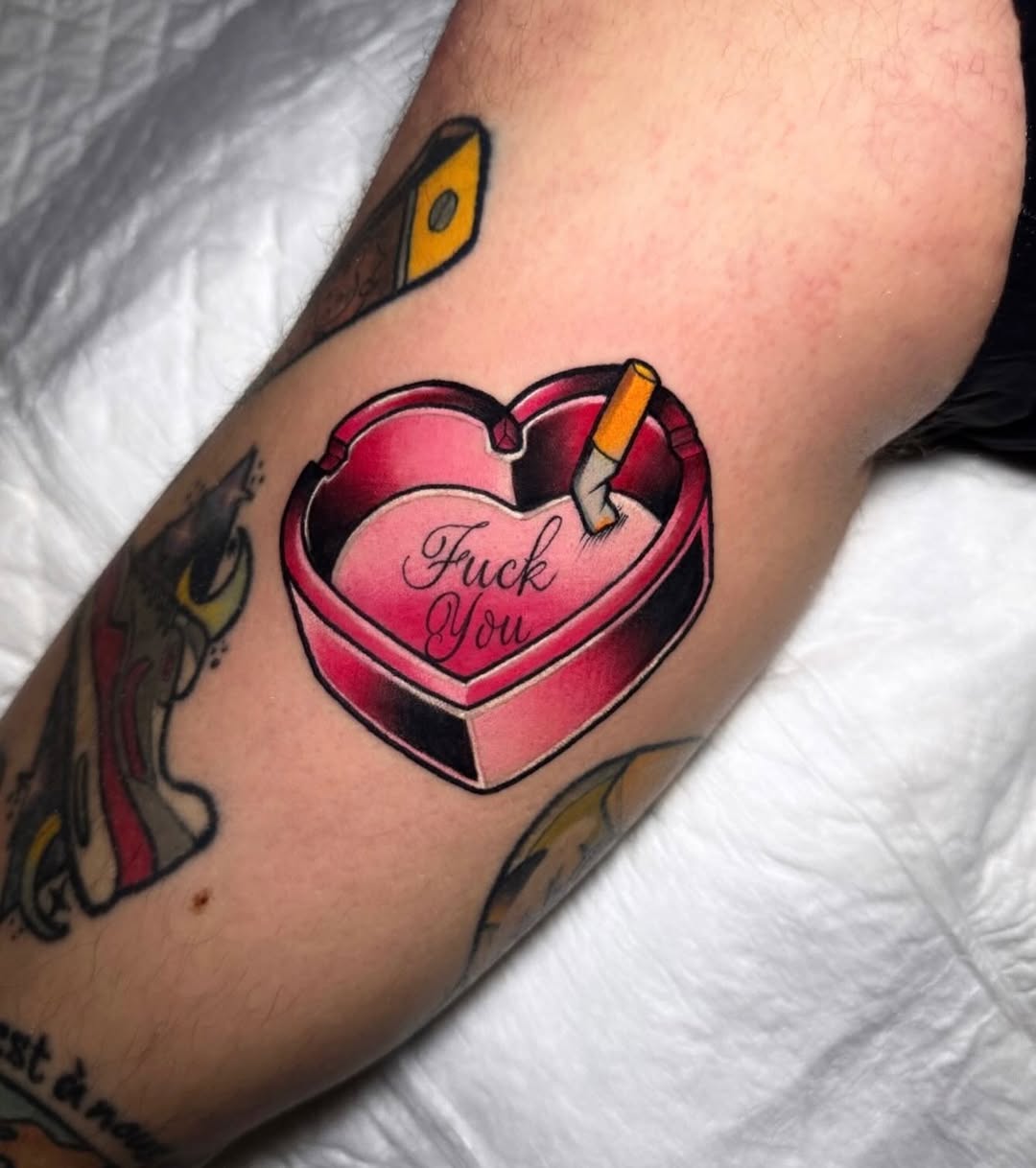 Pink heart-shaped box with cigarette and "Fuck You" text on upper arm, with yellow-eyed bird tattoo visible above.