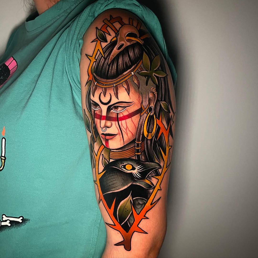 Colorful upper arm tattoo featuring a mystical figure with face markings, long dark hair, bird imagery, and orange flame-like designs.
