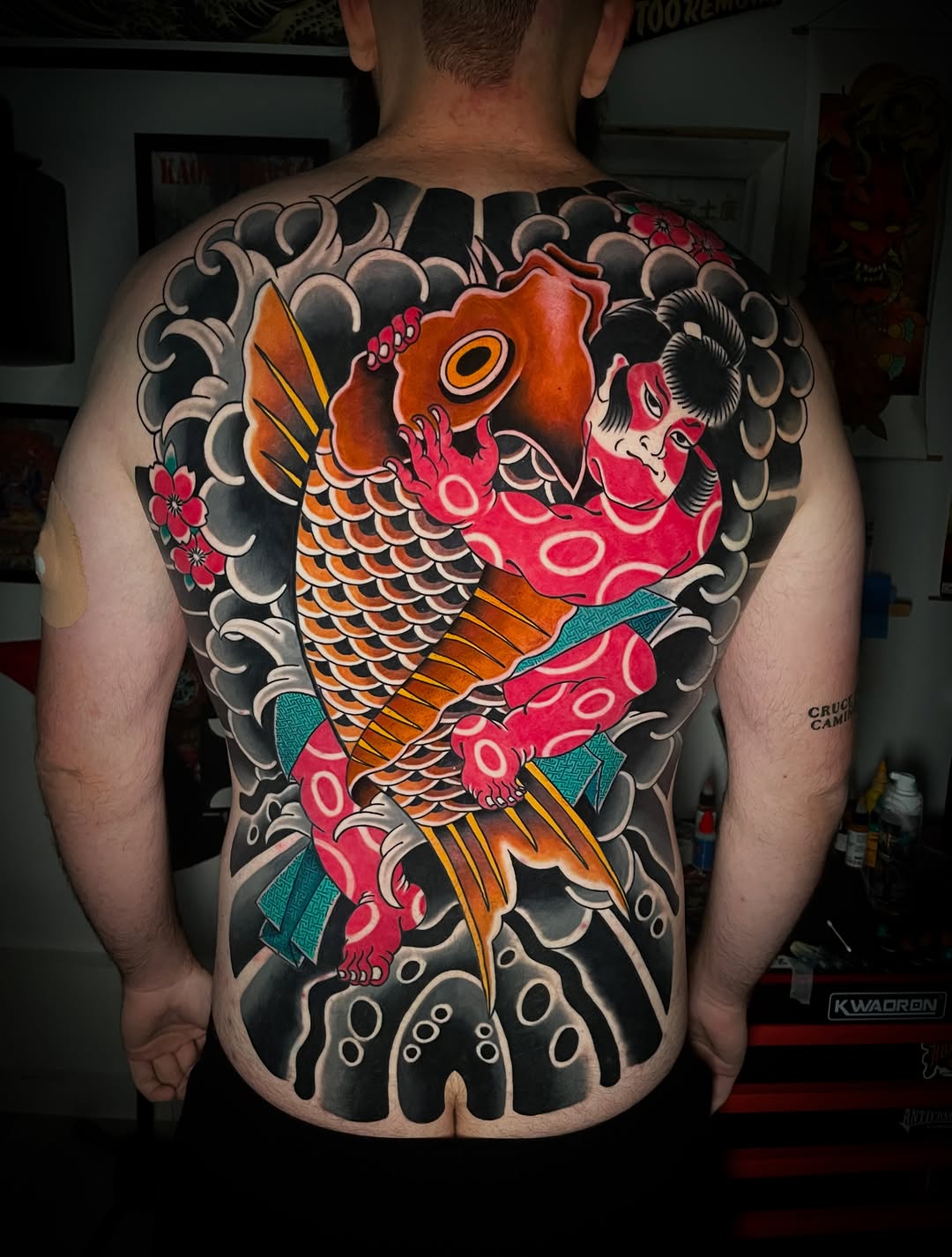 Colorful koi fish tattoo covering the entire back with orange and red scales, surrounded by black cloud motifs and small red flowers.