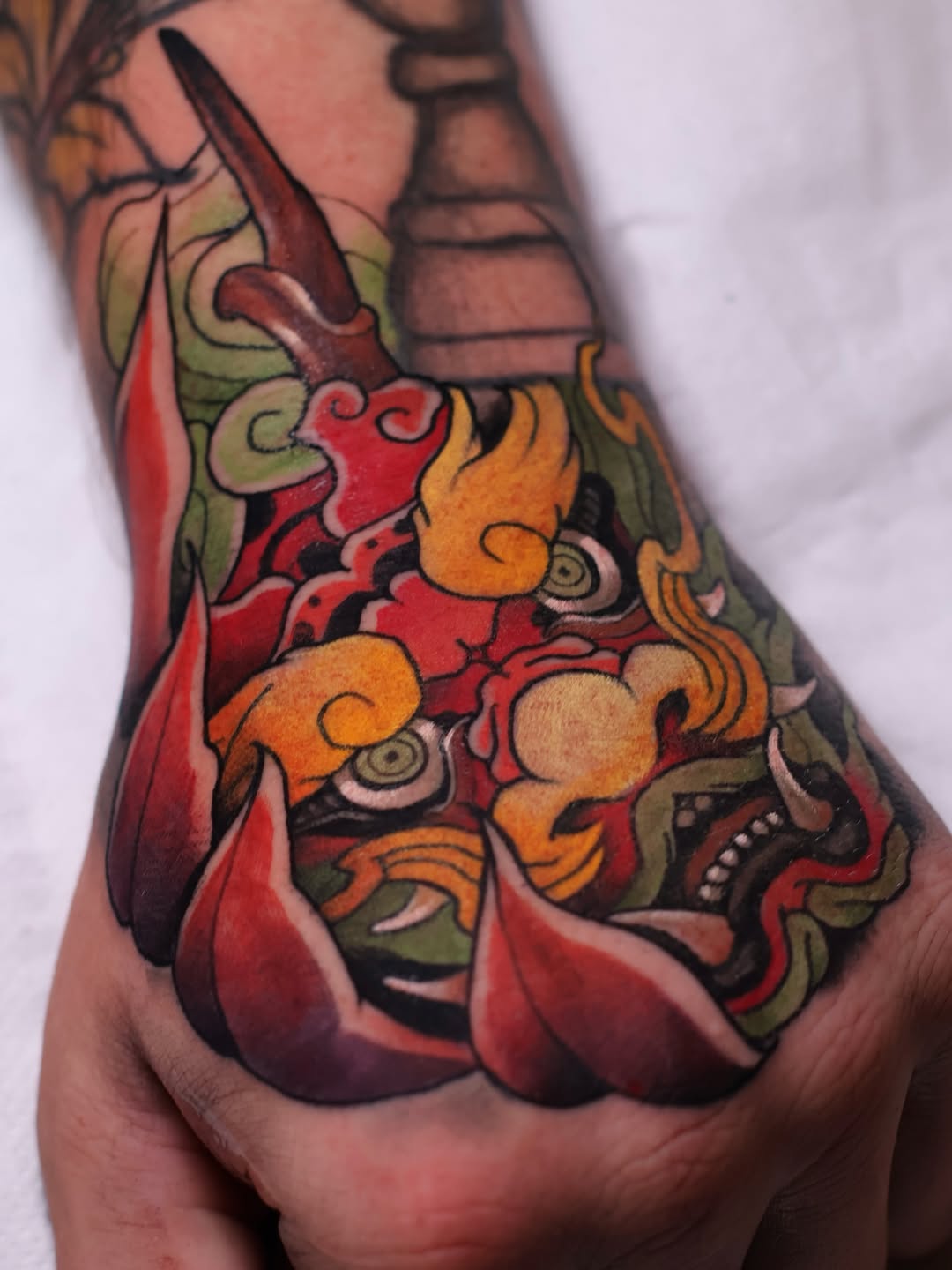 Colorful hand tattoo featuring ornamental creatures with orange, red, and green details, intricate scrollwork, and decorative elements on the knuckles and fingers.