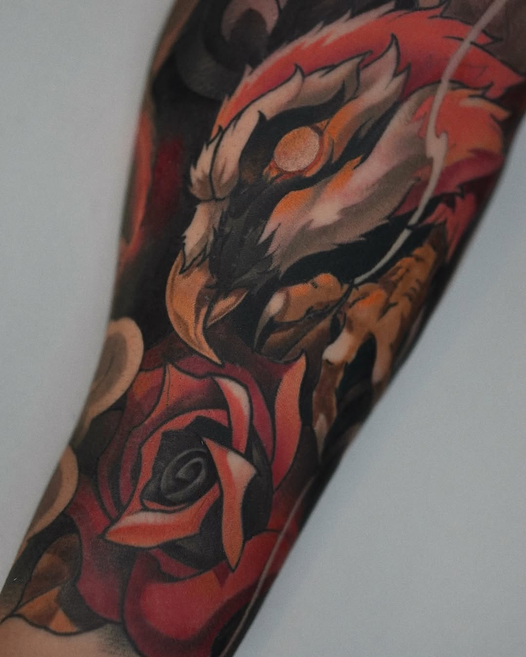 Colorful arm tattoo featuring a demon face with orange eyes and red mouth above a large red rose with black details and shading.