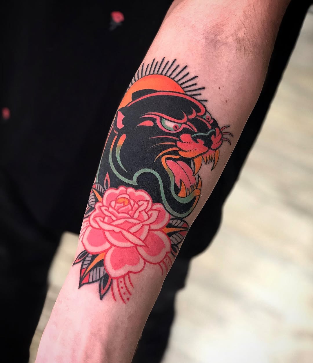 Colorful arm tattoo featuring a black panther head with orange sun halo above a pink rose, located on the forearm.