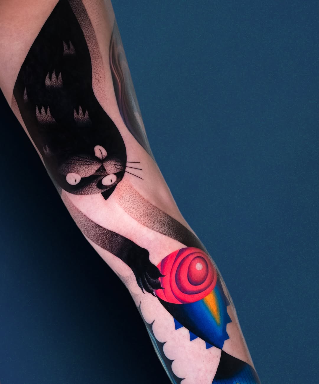 Arm tattoo featuring a cat's face with three eyes and whiskers, a red spiral lollipop, and a rocket with blue and yellow details against skin.