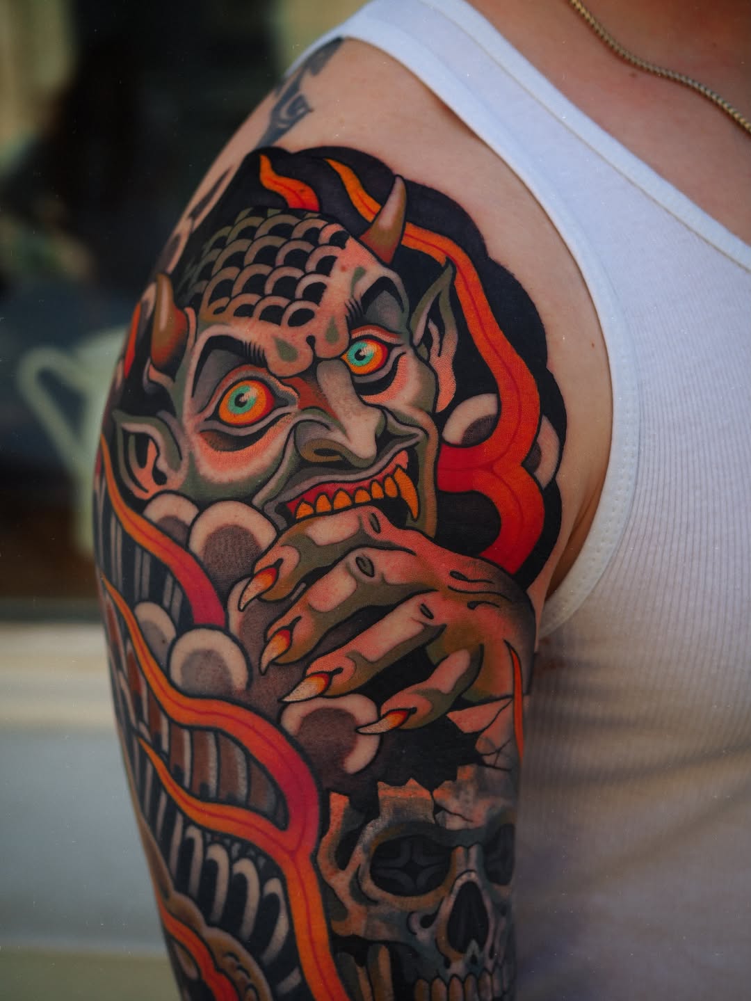 Colorful demon or oni mask tattoo on upper arm featuring red eyes, orange and black details, with a skull visible below, displayed on pale skin.