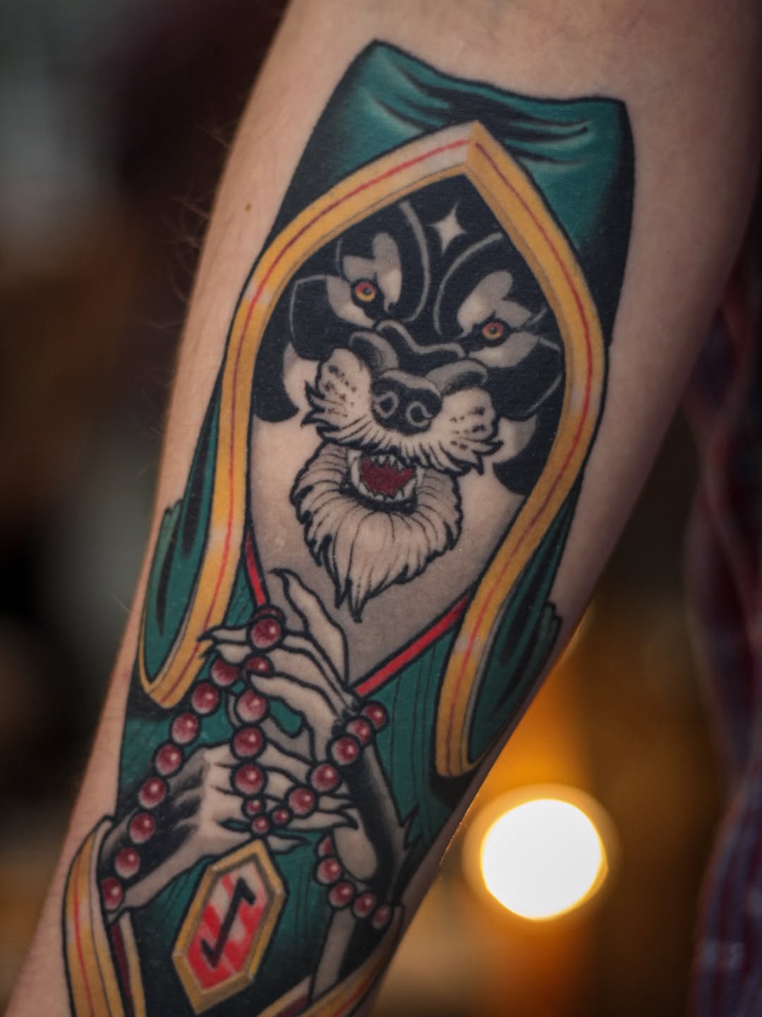Coffin-shaped tattoo featuring a skeletal face with red eyes, wearing a green cloak and holding red beads, framed in gold and yellow borders.