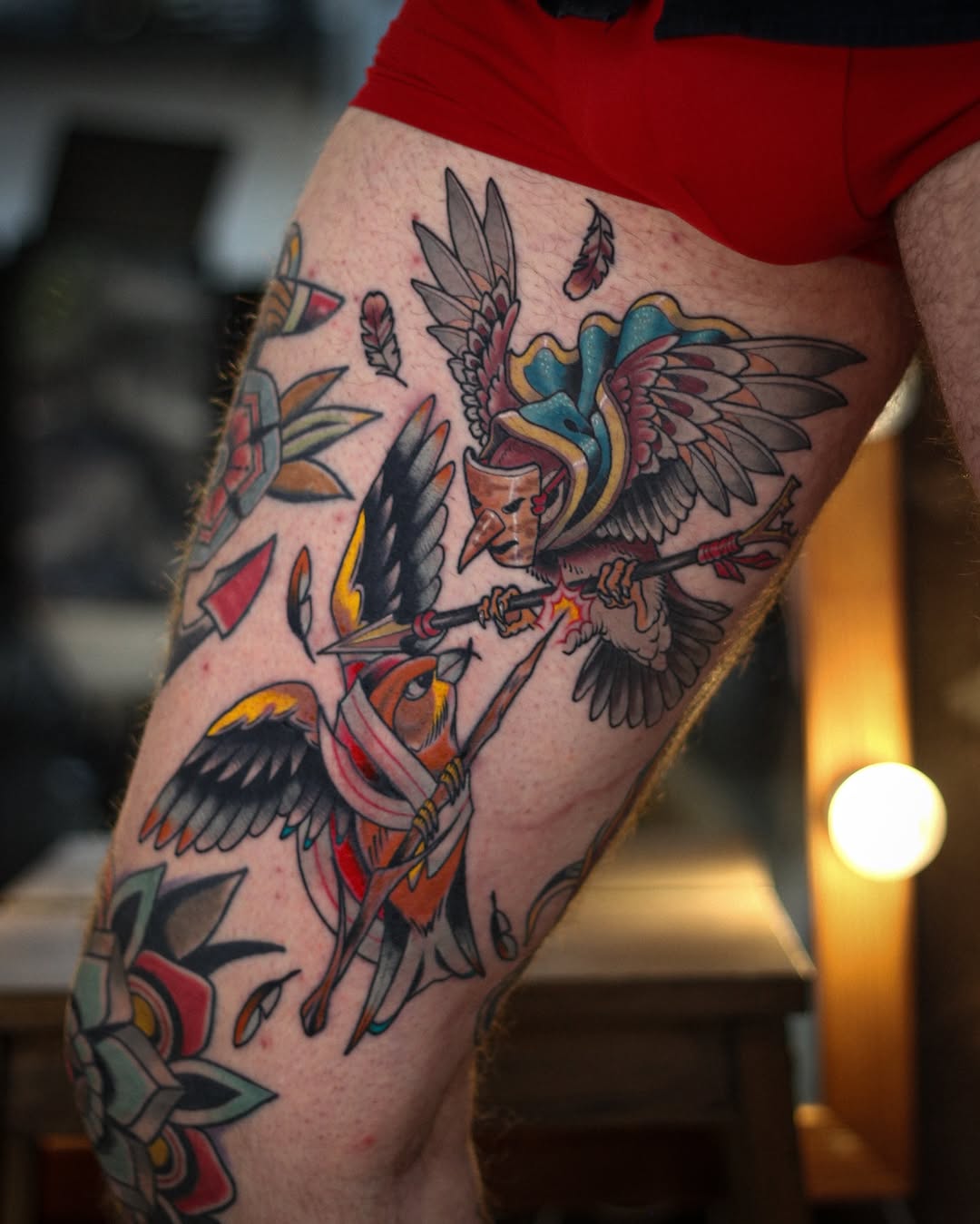 Colorful winged figures with feathers and flowers tattooed on a man's thigh, featuring red, blue, yellow, and black ink.