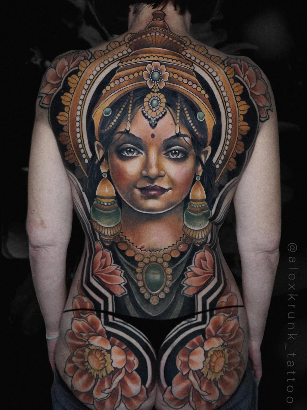 Back piece tattoo featuring a woman's face with ornate jewelry, decorative headdress, and lotus flowers in orange, gold, and black tones.