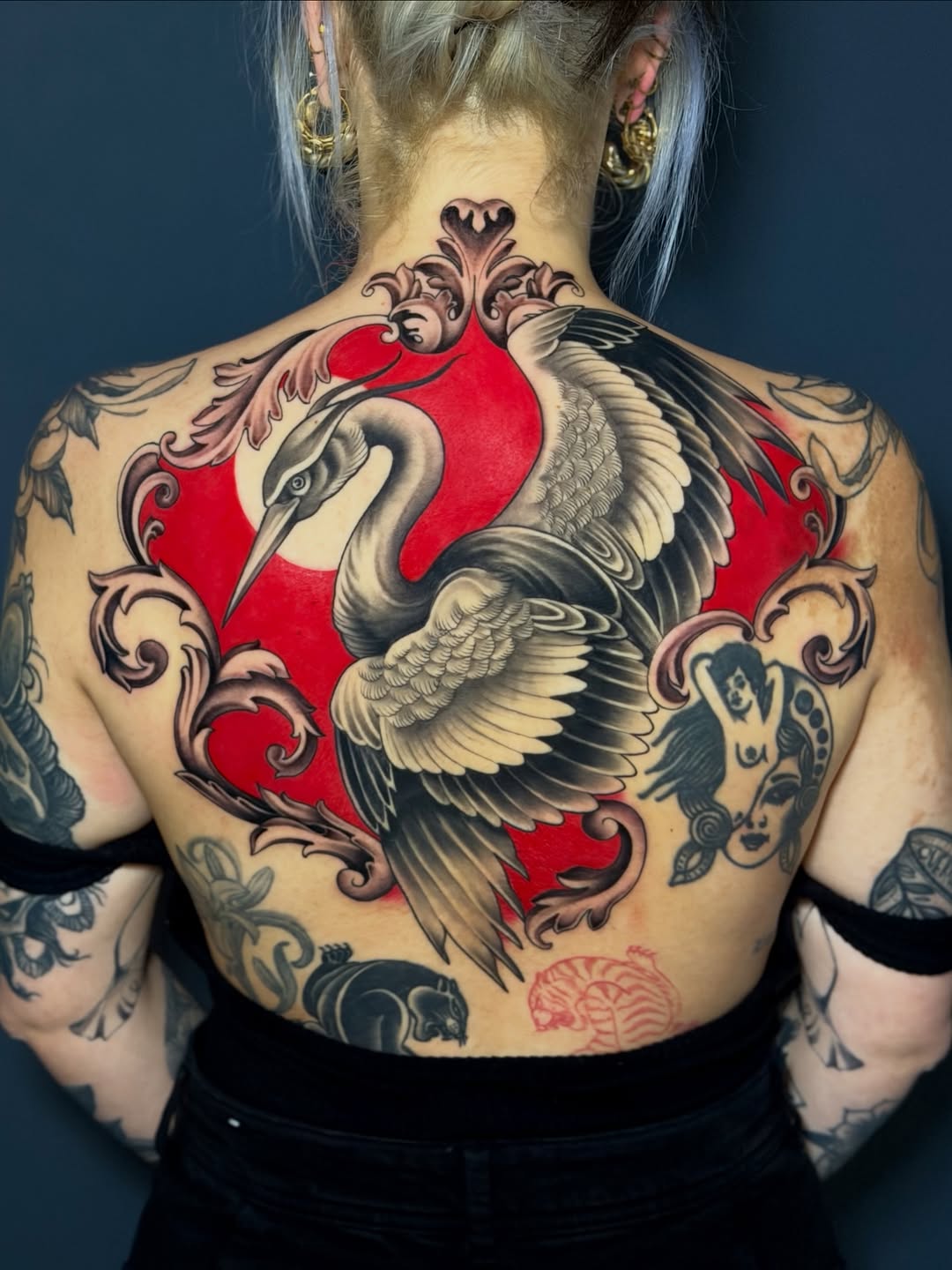 Back tattoo featuring a crane with spread wings centered on a red shield, surrounded by ornamental scrollwork in black, red, and grey ink.