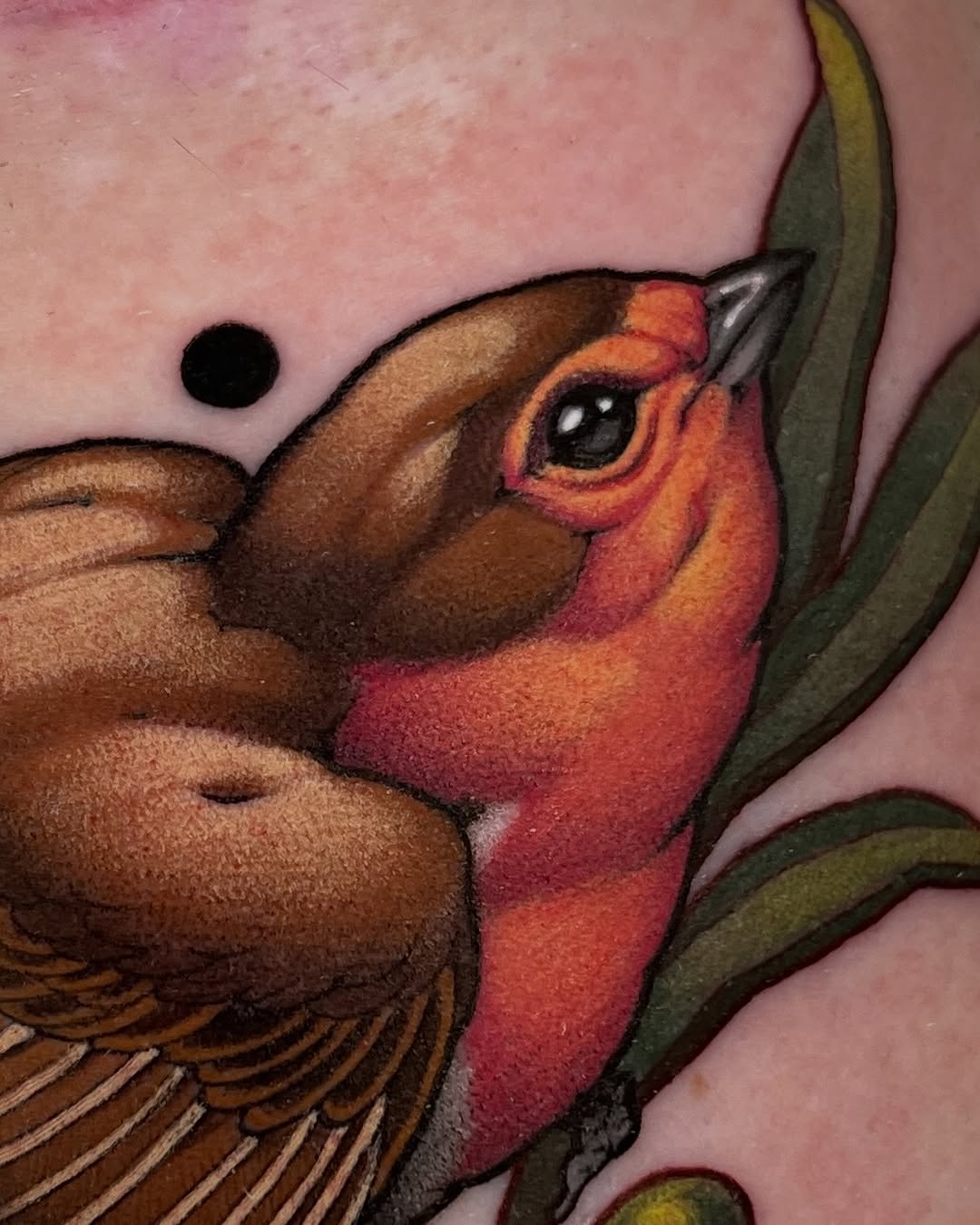 Colorful bird tattoo with brown wings, red and orange head, and green leaf details on pink skin.