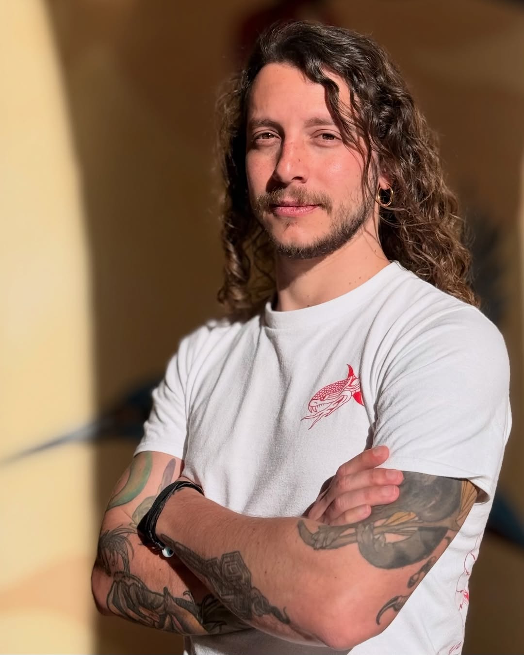 Man with long brown hair and facial hair wearing a white t-shirt displaying multiple arm tattoos including birds and geometric designs in various colors.