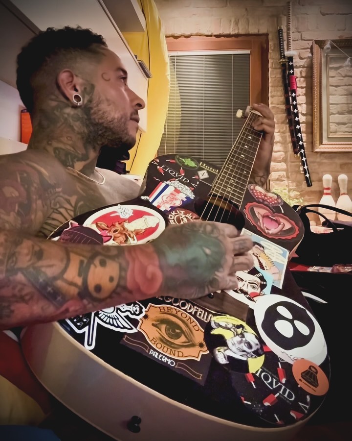 Shirtless man with extensive colorful chest and arm tattoos holds a decorated electric guitar while seated indoors.