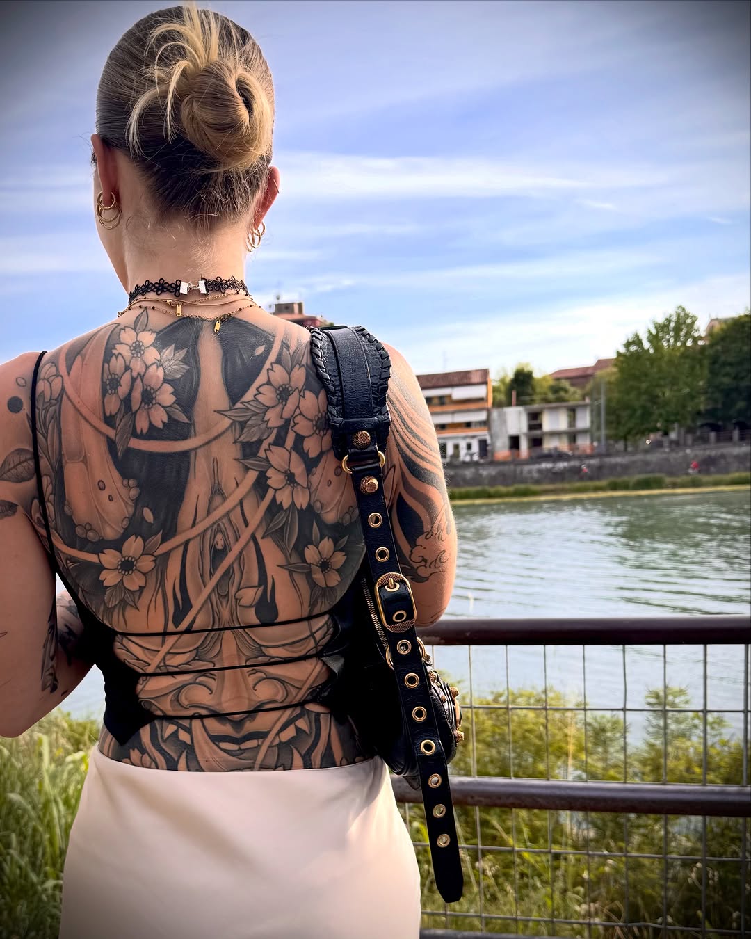 Woman displaying large-scale floral and botanical back tattoo in black ink with detailed linework, photographed outdoors by a waterway.