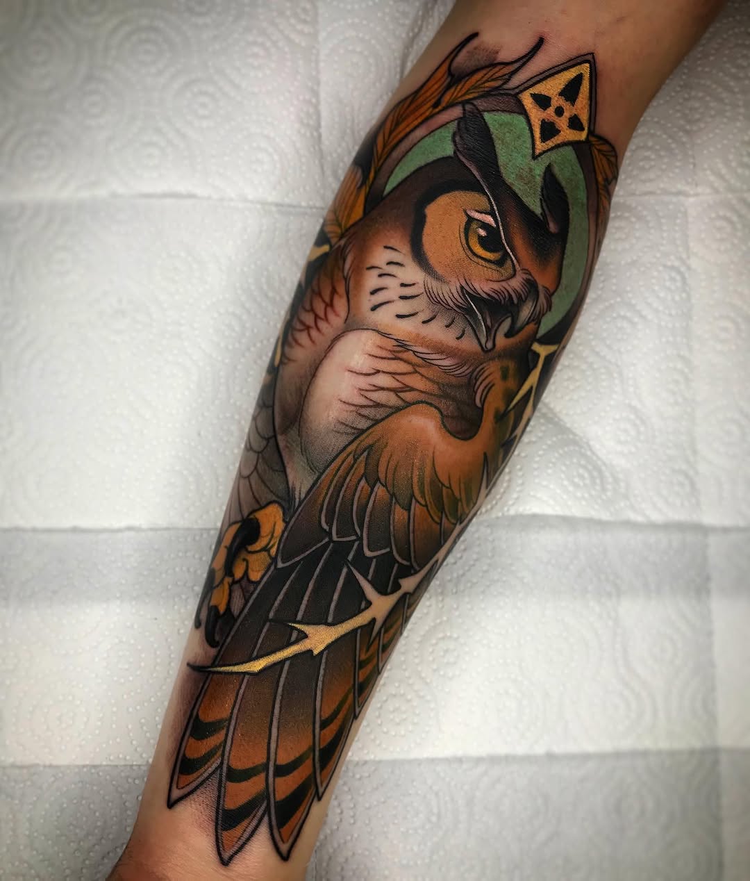 Detailed owl tattoo on a forearm featuring brown and orange plumage, green headpiece, yellow eyes, and geometric crown, displayed against a white textured background.