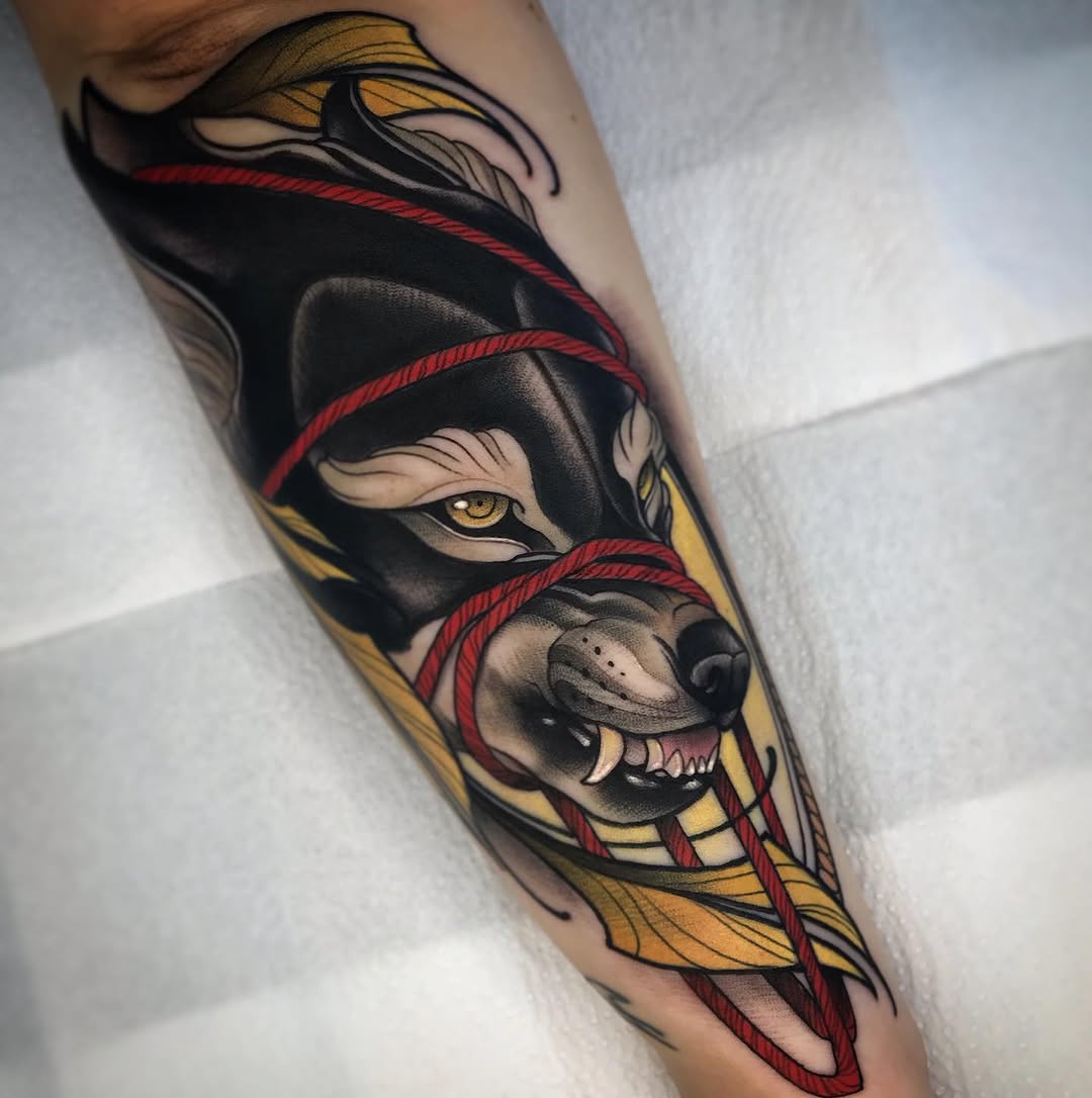 Colorful arm tattoo featuring a masked face with golden wings and red rope details wrapped around black and yellow geometric sections.