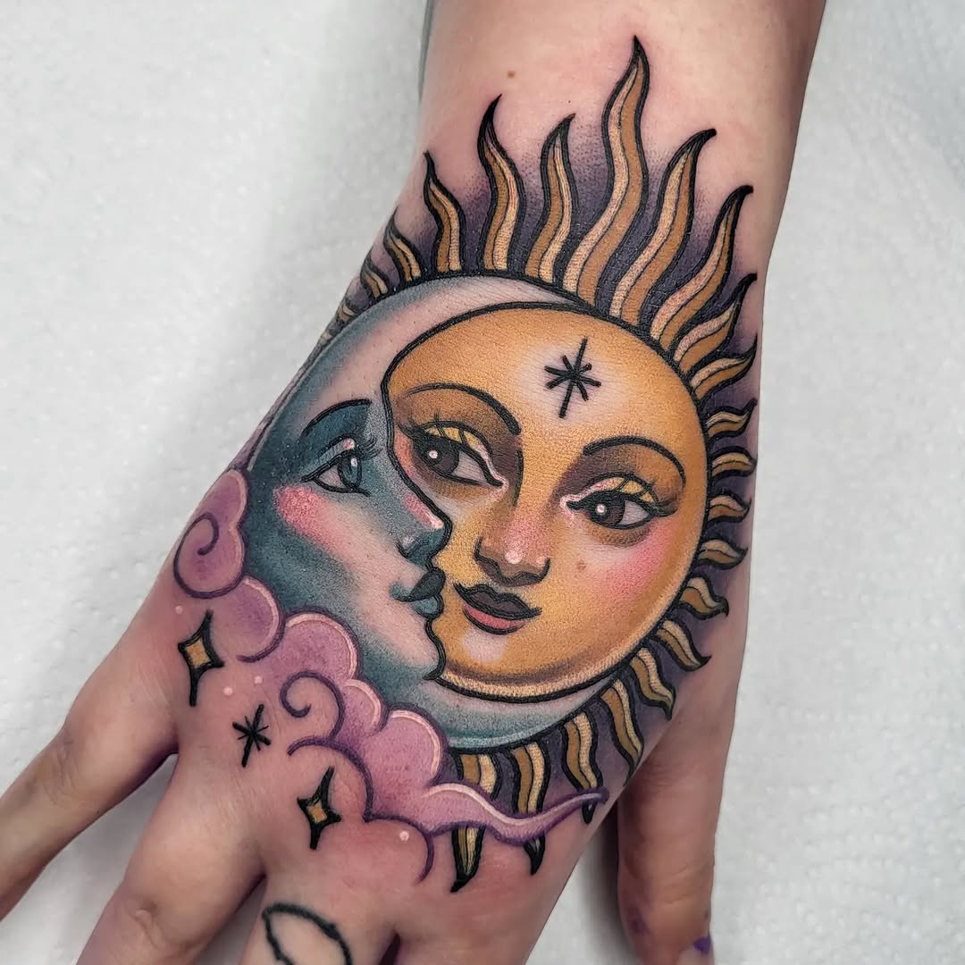 Celestial sun and moon face tattoo on hand with gold, purple, and teal coloring, featuring flames, clouds, and decorative stars.