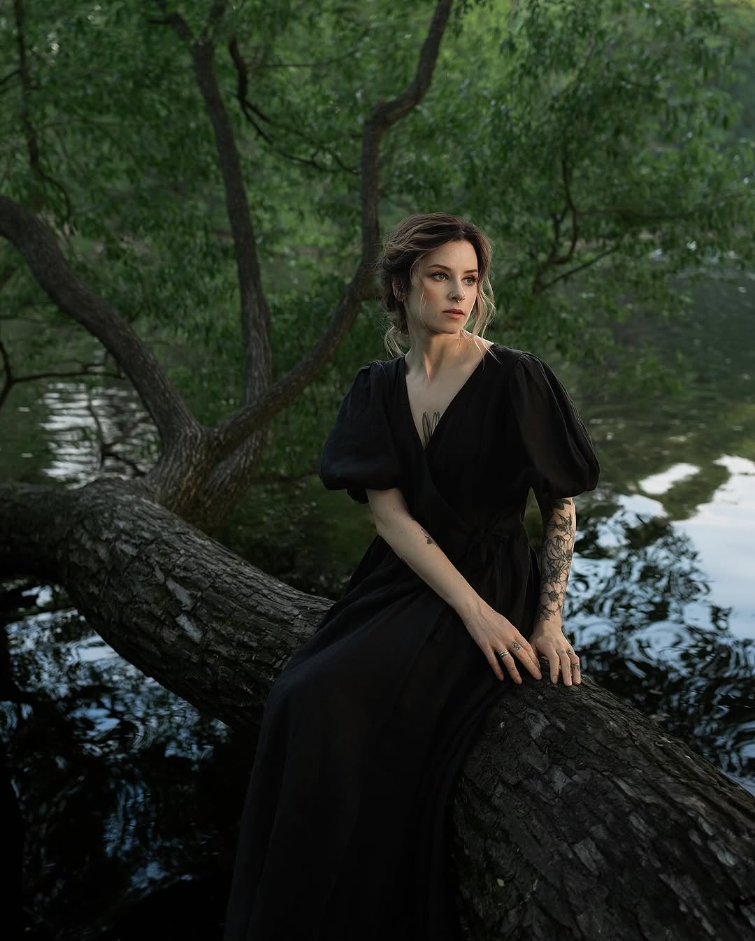 Woman wearing black dress seated on tree branch over water displays an ornate floral tattoo on her right forearm in dark ink.