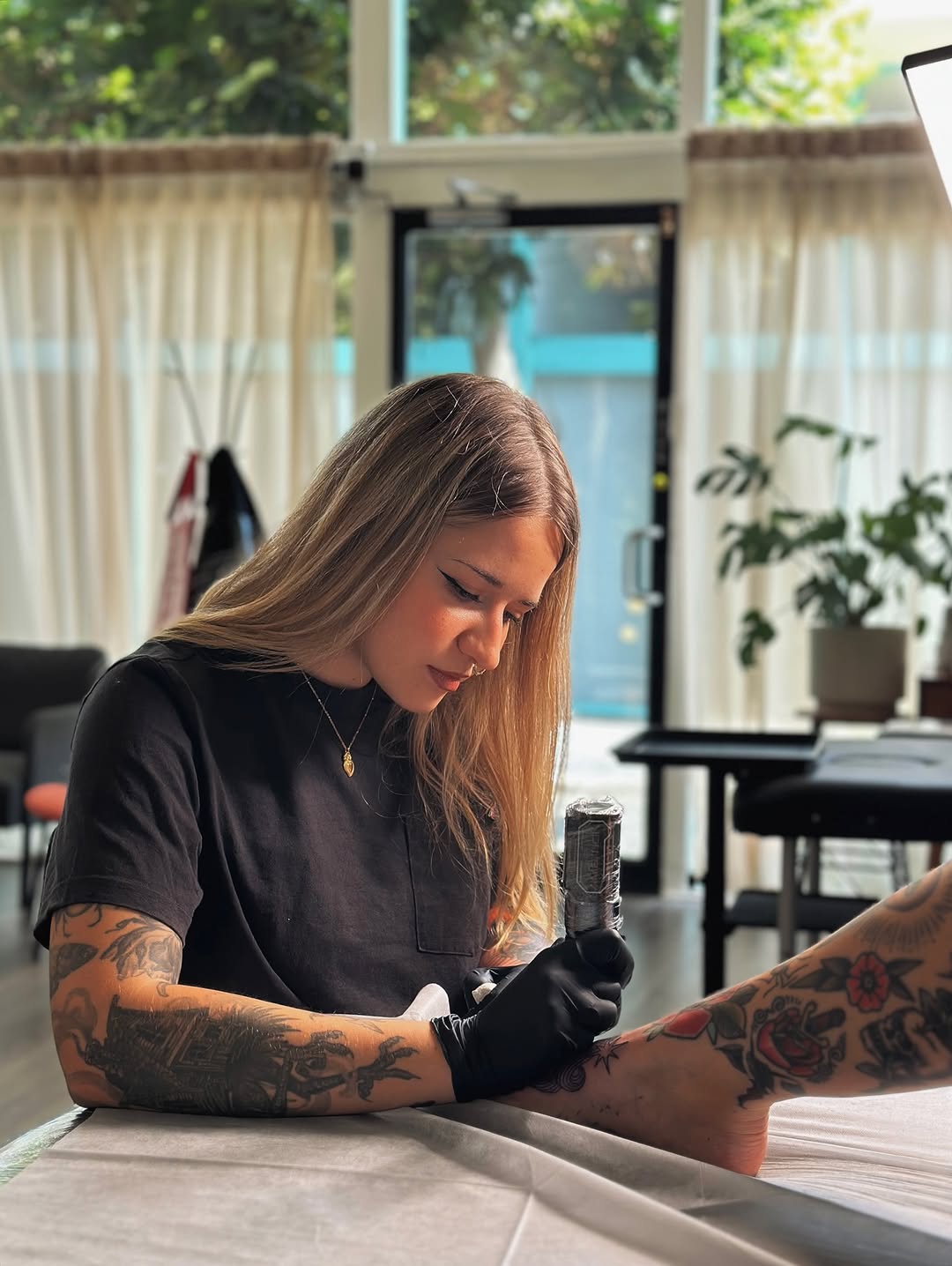 Tattoo artist wearing black gloves applies red rose tattoo to client's arm in bright studio with glass doors and greenery.