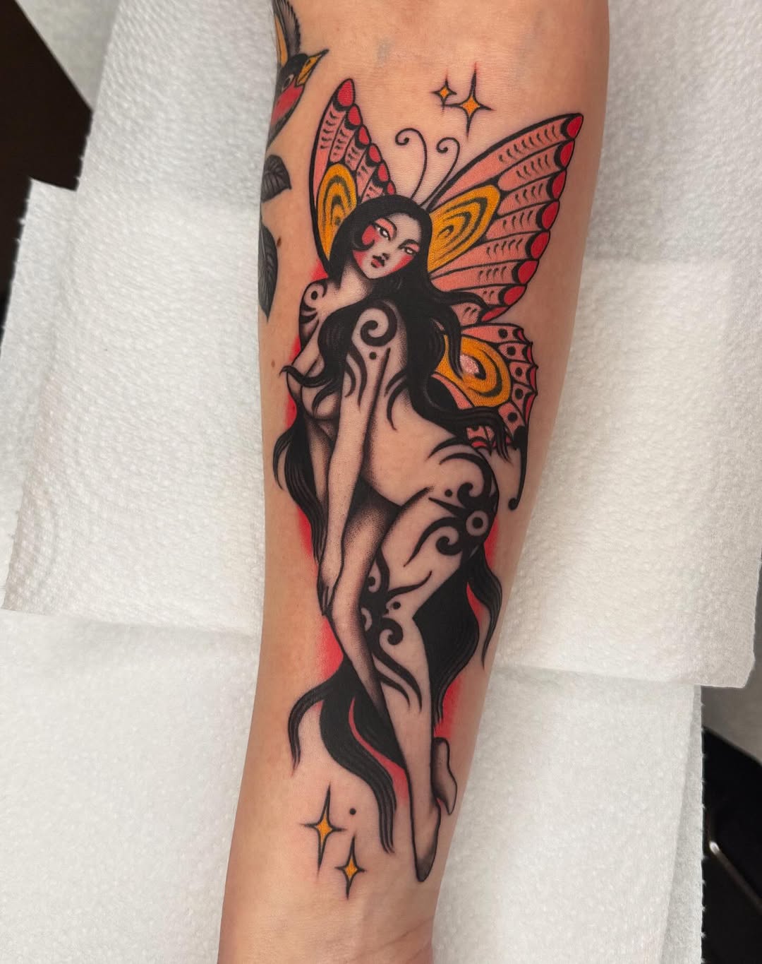 Fairy woman with butterfly wings in red, gold, and black ink on forearm, featuring decorative swirls and small stars.