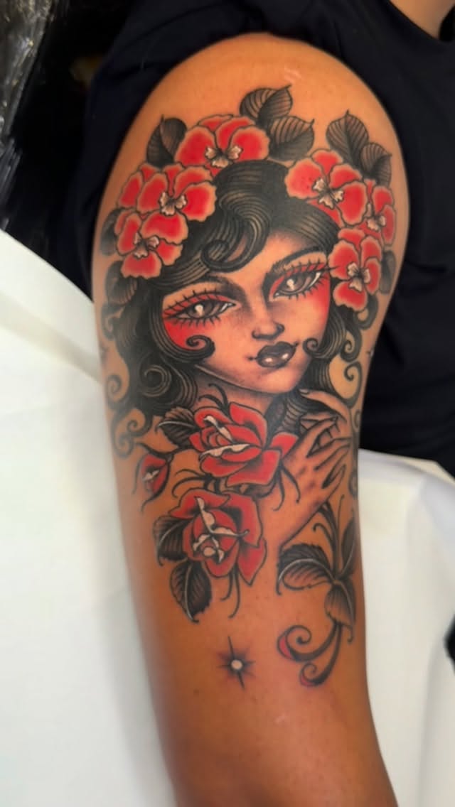 Colorful shoulder tattoo featuring a woman's face with red flowers in her hair, red facial markings, and red roses on her chest and arm with decorative scrollwork.