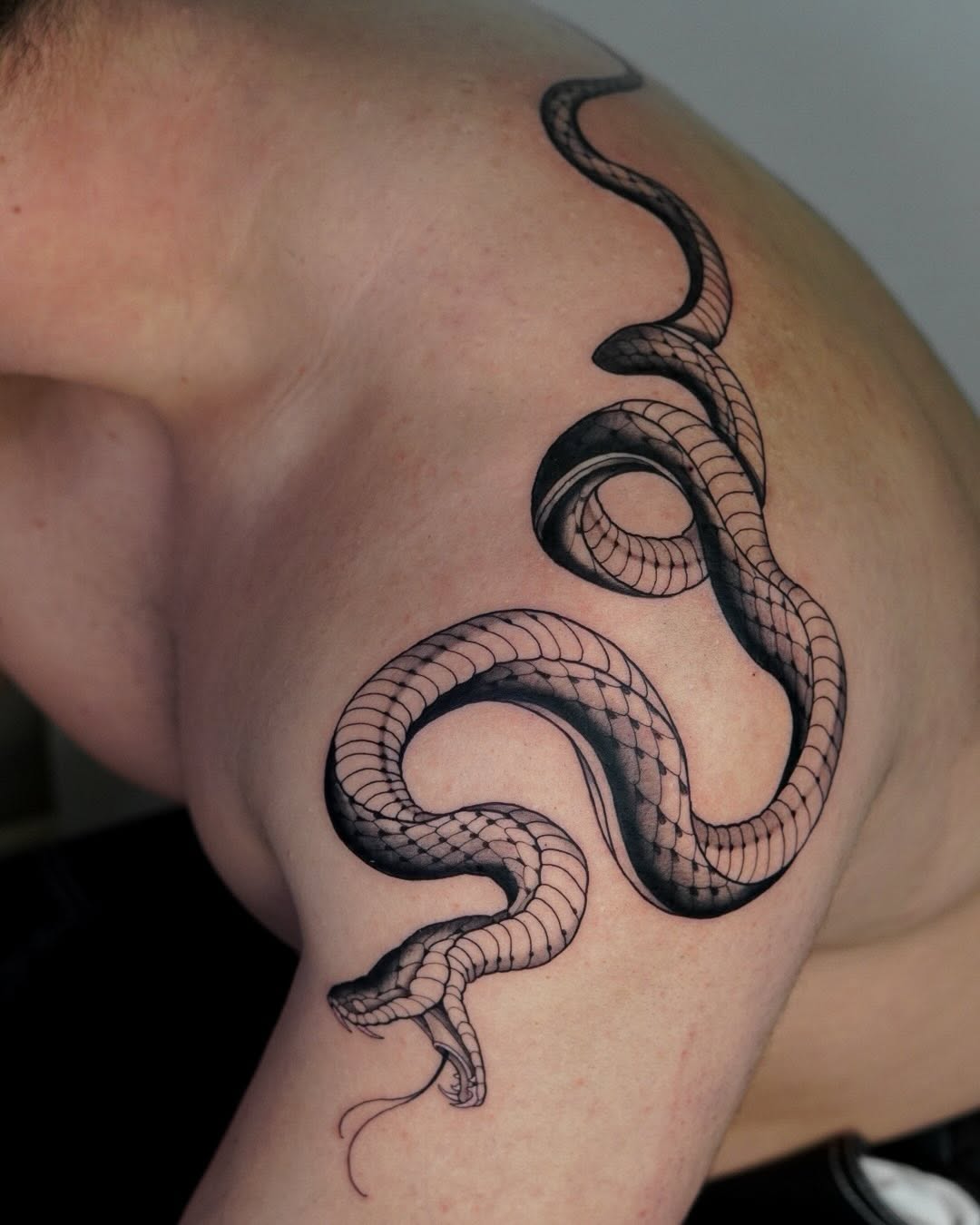 Detailed black snake tattoo with scaled texture wrapping around the upper arm and shoulder of a person, featuring two coiled sections with an open mouth.