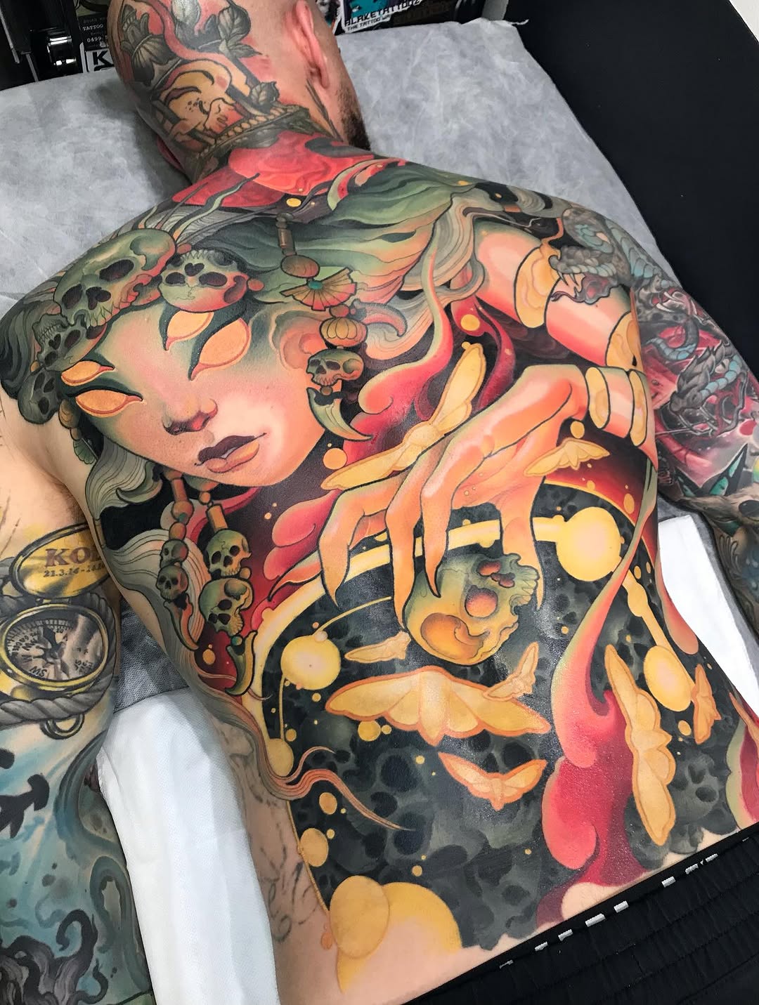 Large-scale chest and shoulder tattoo featuring a woman's face with ornamental headdress, skulls, flowers, and decorative elements in red, yellow, orange, green, and black.