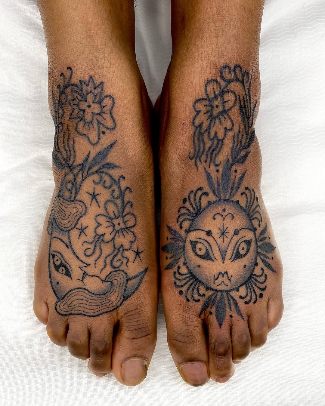 Matching symmetrical foot tattoos featuring celestial faces and floral motifs in dark blue ink on brown skin.