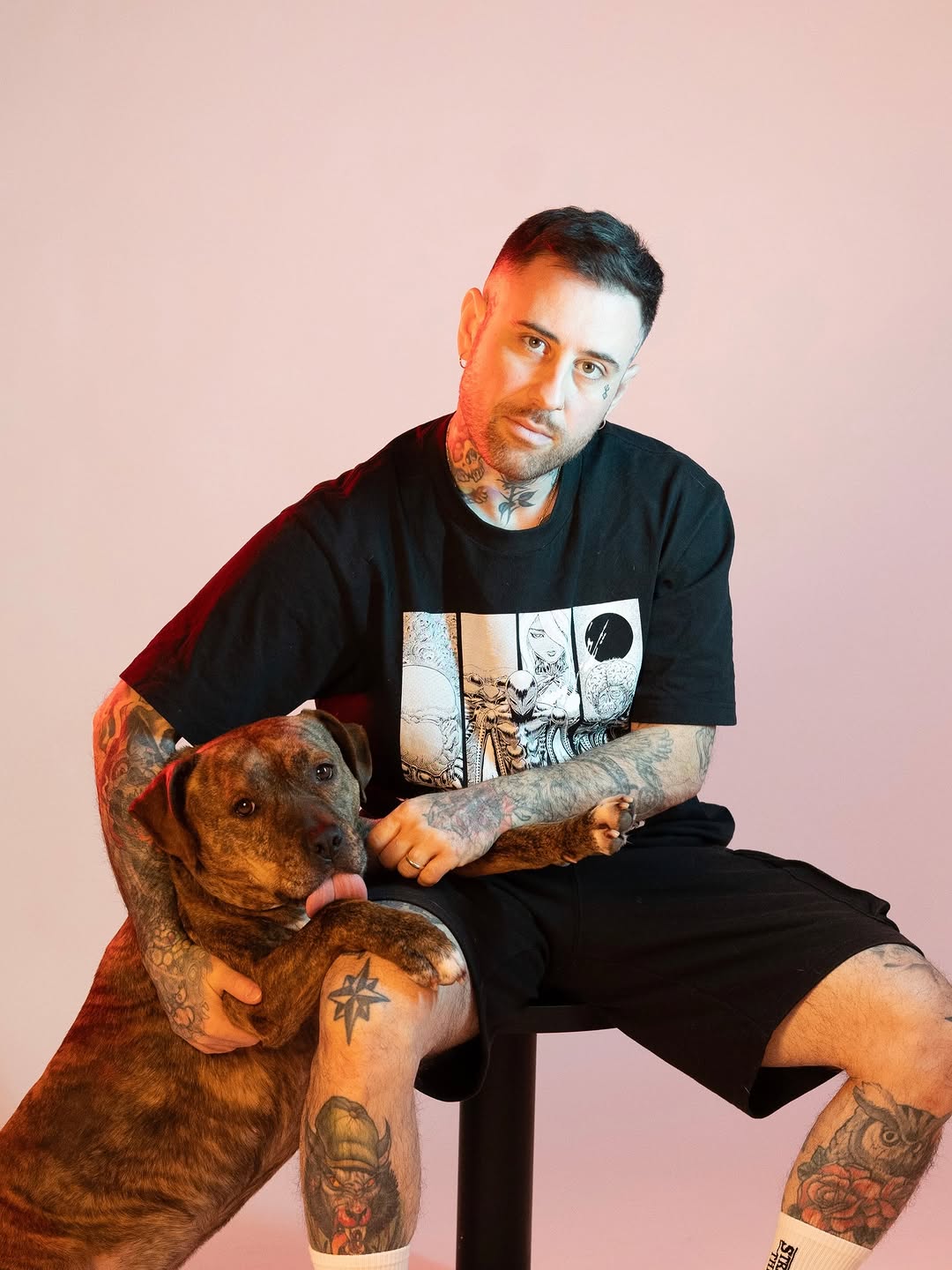 Tattooed man with brown dog posing against pink background, displaying extensive black ink on neck, arms, and legs in portfolio photograph.
