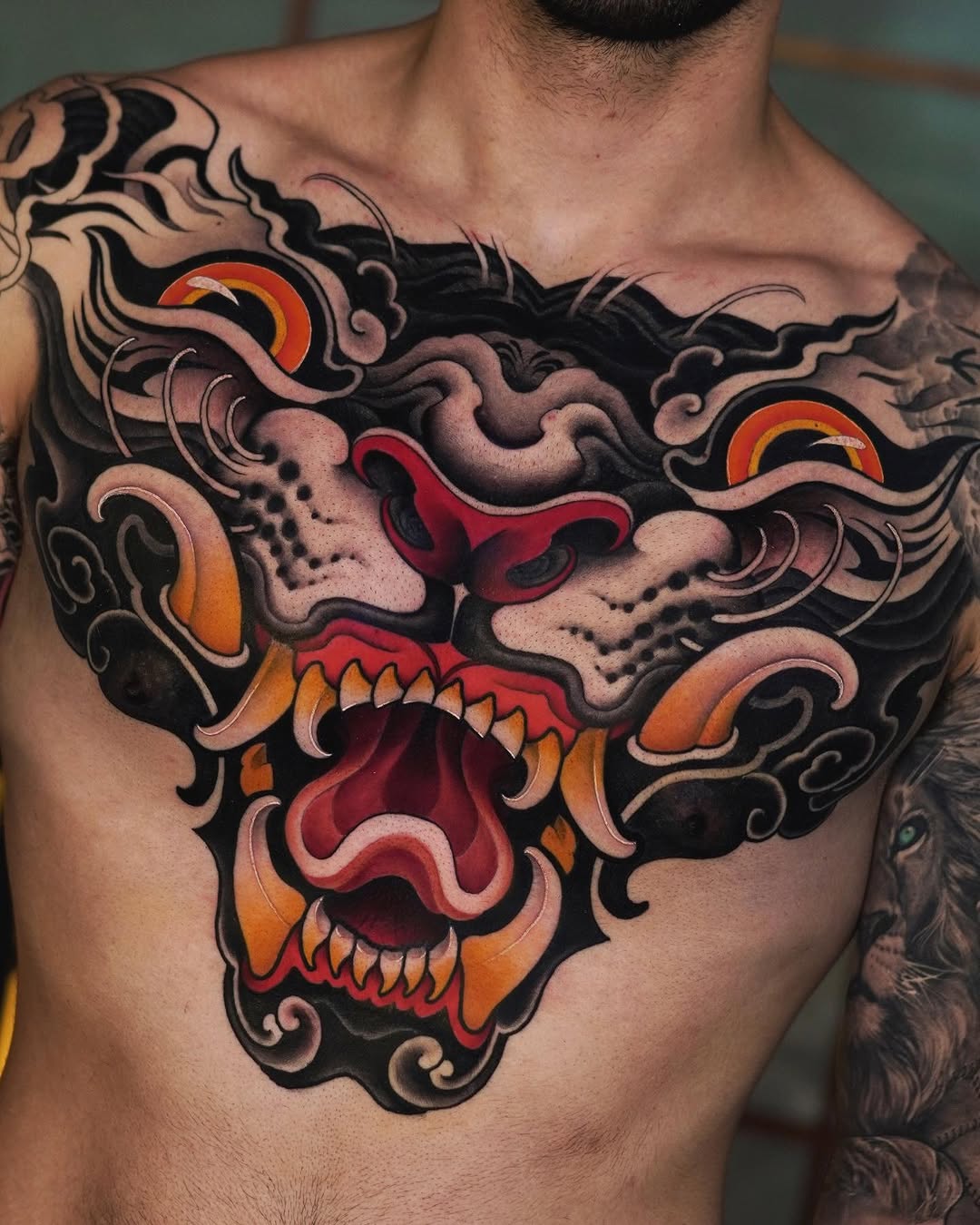 Large colorful chest tattoo featuring a demon face with red and orange accents, ornate black scrollwork, and skull imagery on a man's torso.