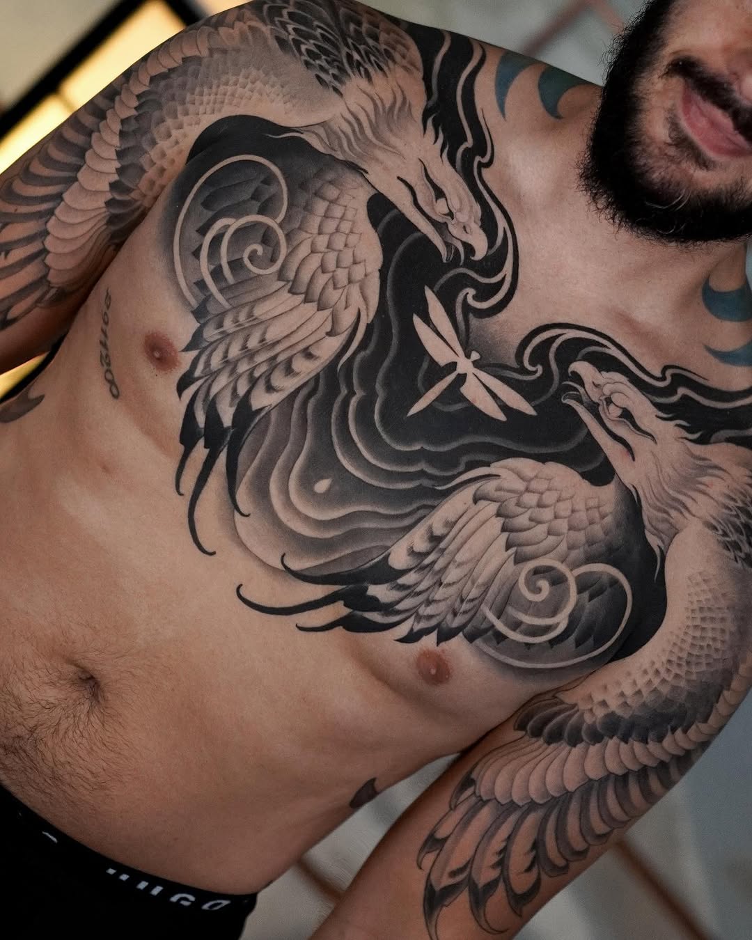 Large-scale chest and shoulder tattoo featuring winged mythological creatures with intricate feather details and spiral ornaments in black and gray ink.