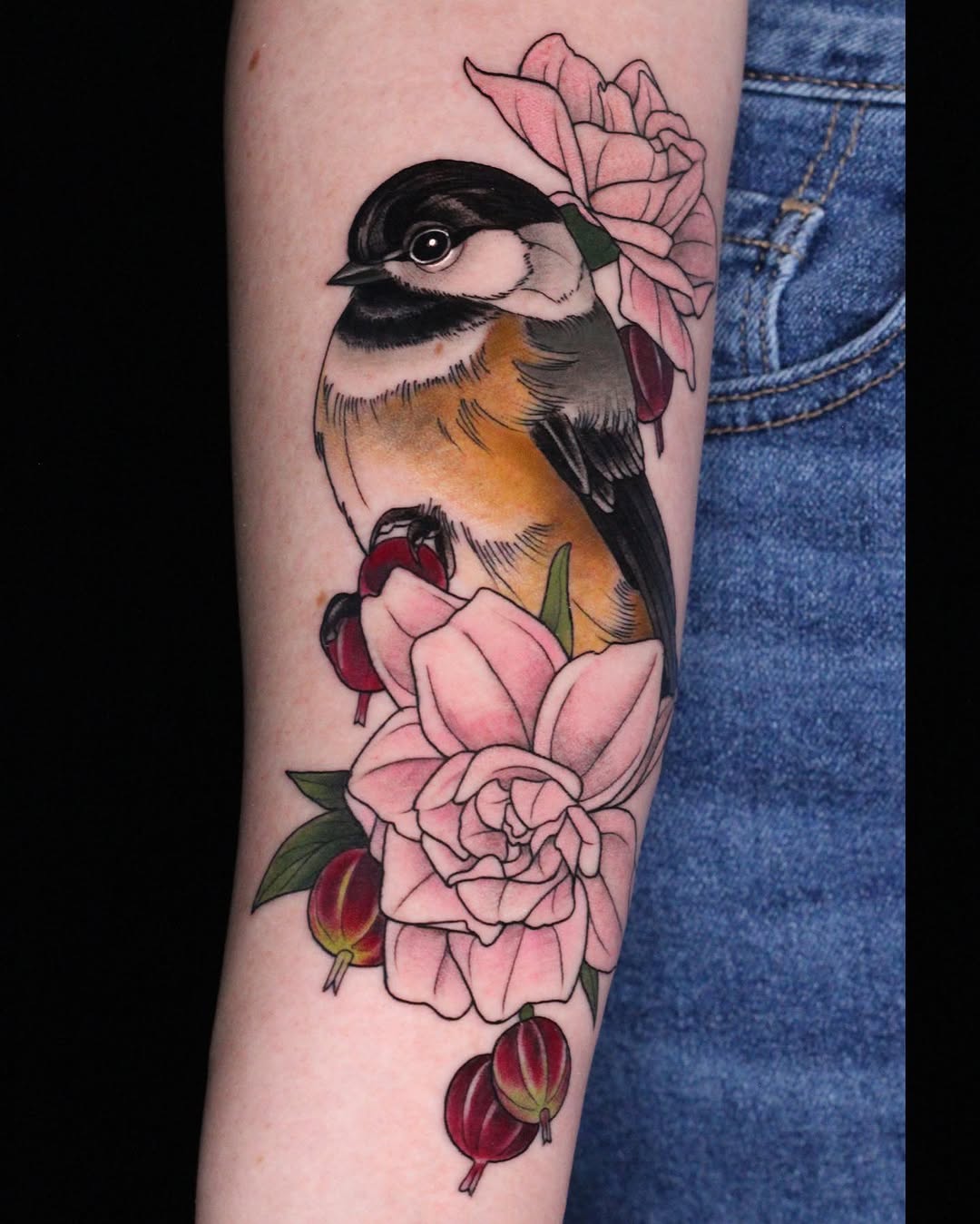 Colorful bird with orange and black plumage perched among pink roses and red berries tattooed on the forearm.