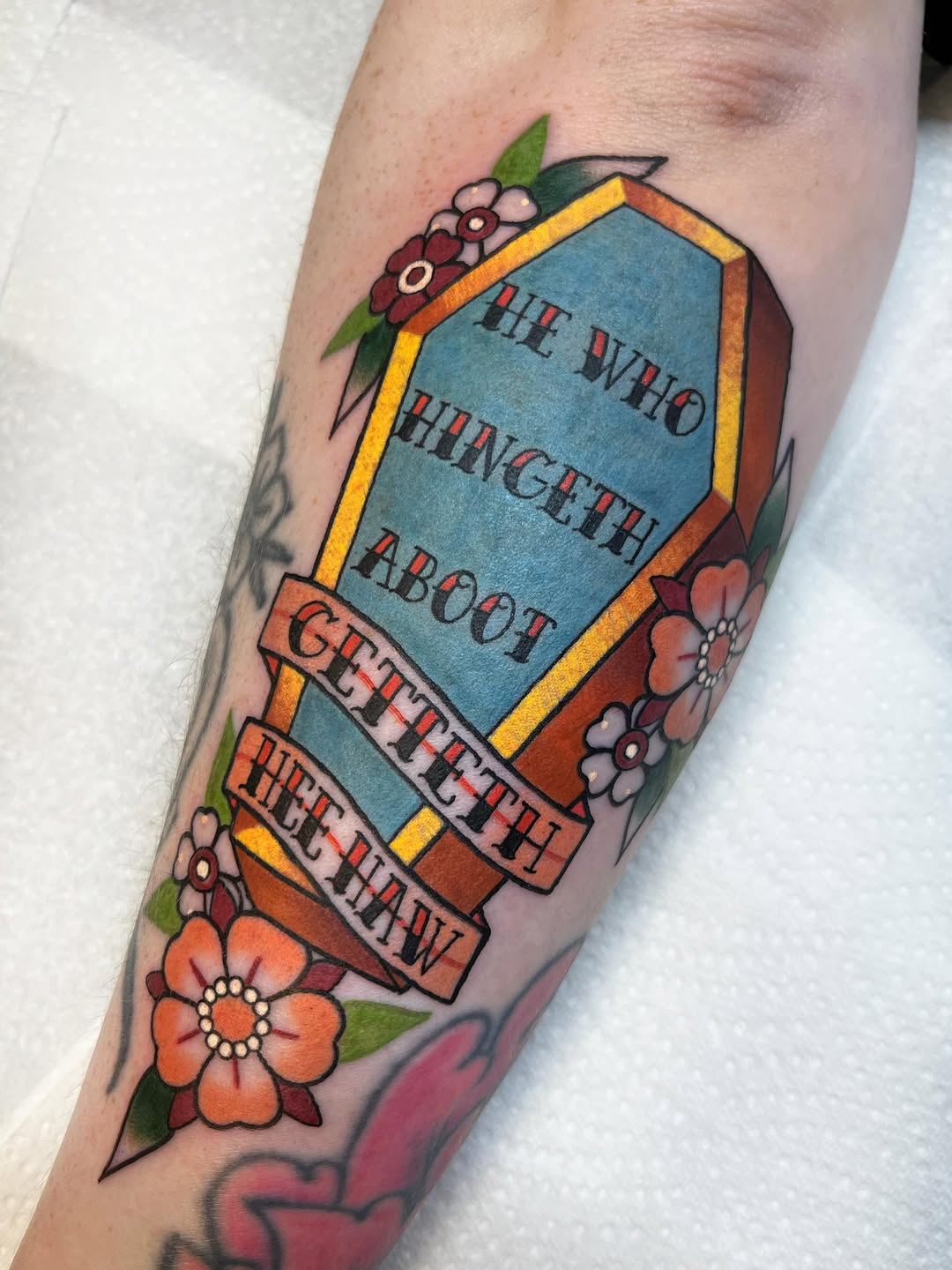 Coffin-shaped tattoo with blue center panel reading "He Who Hingeth About Getteth The Heck Outta Here," surrounded by colorful flowers on the calf.