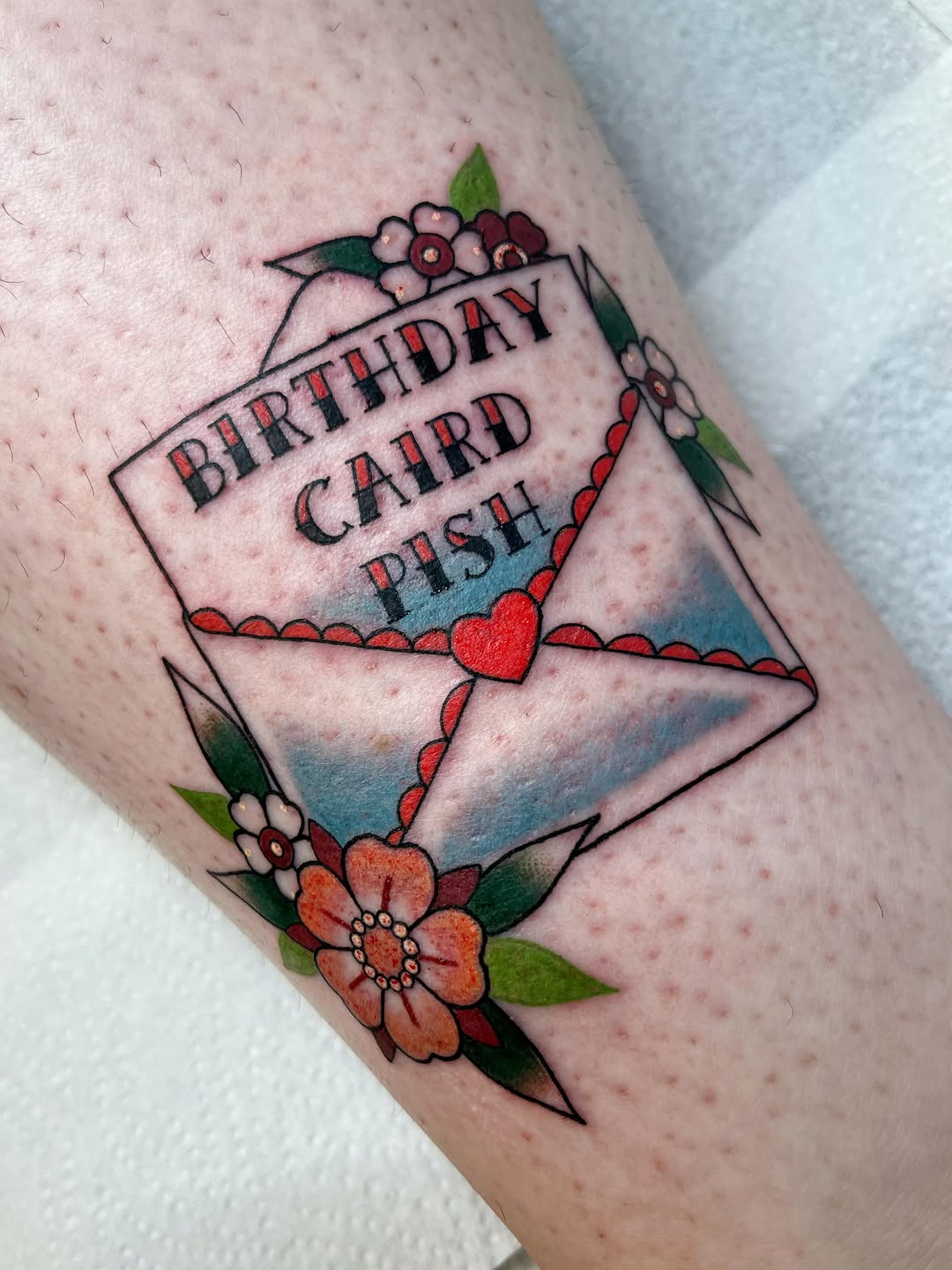 Birthday card push envelope tattoo on an arm featuring flowers, a red heart, blue watercolor wash, and decorative scalloped borders in red, green, and orange.