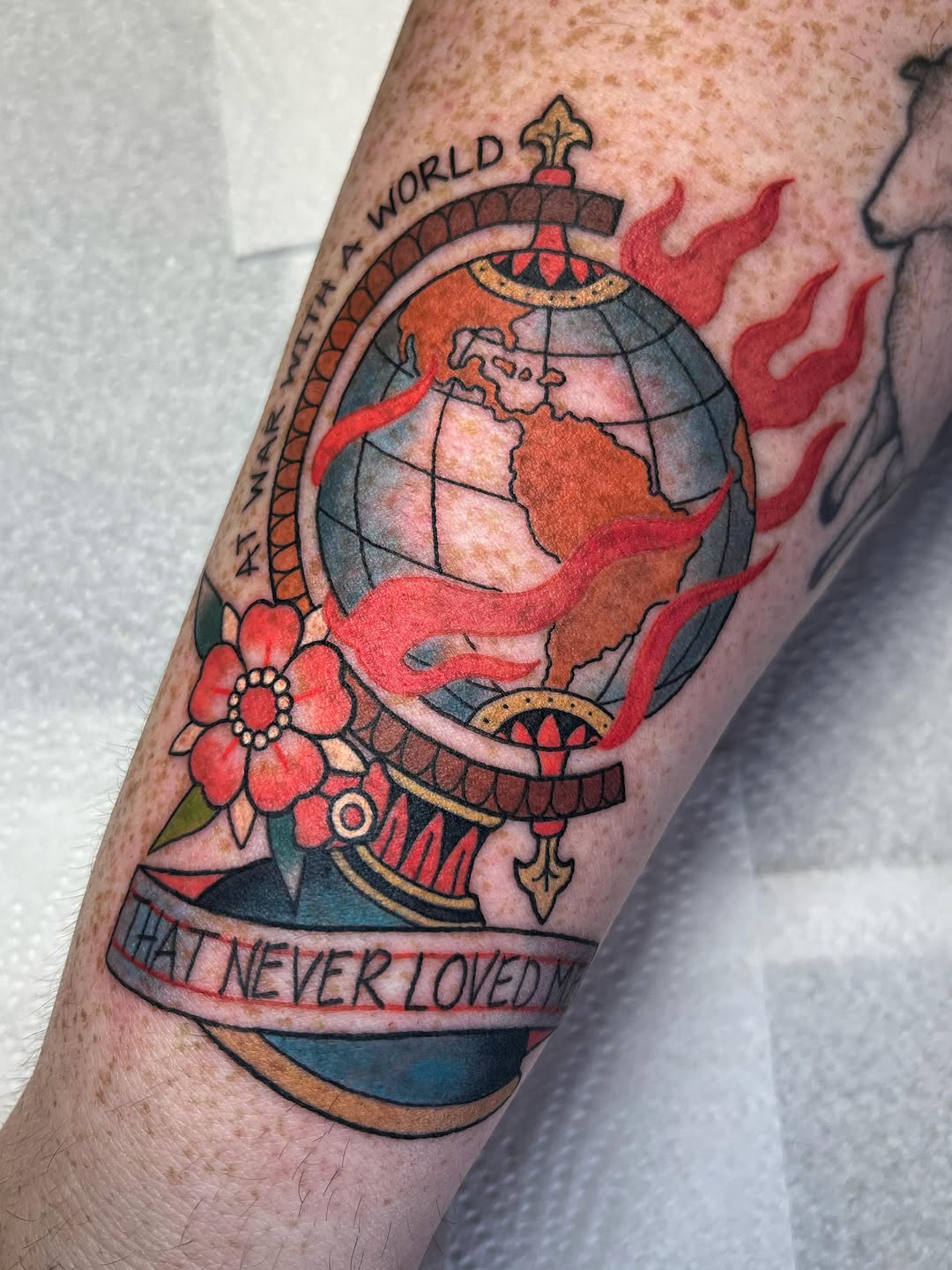Ornate globe tattoo on forearm featuring red flames, a cross top, decorative flower, and text banner reading "what never loved me" in muted earth tones.