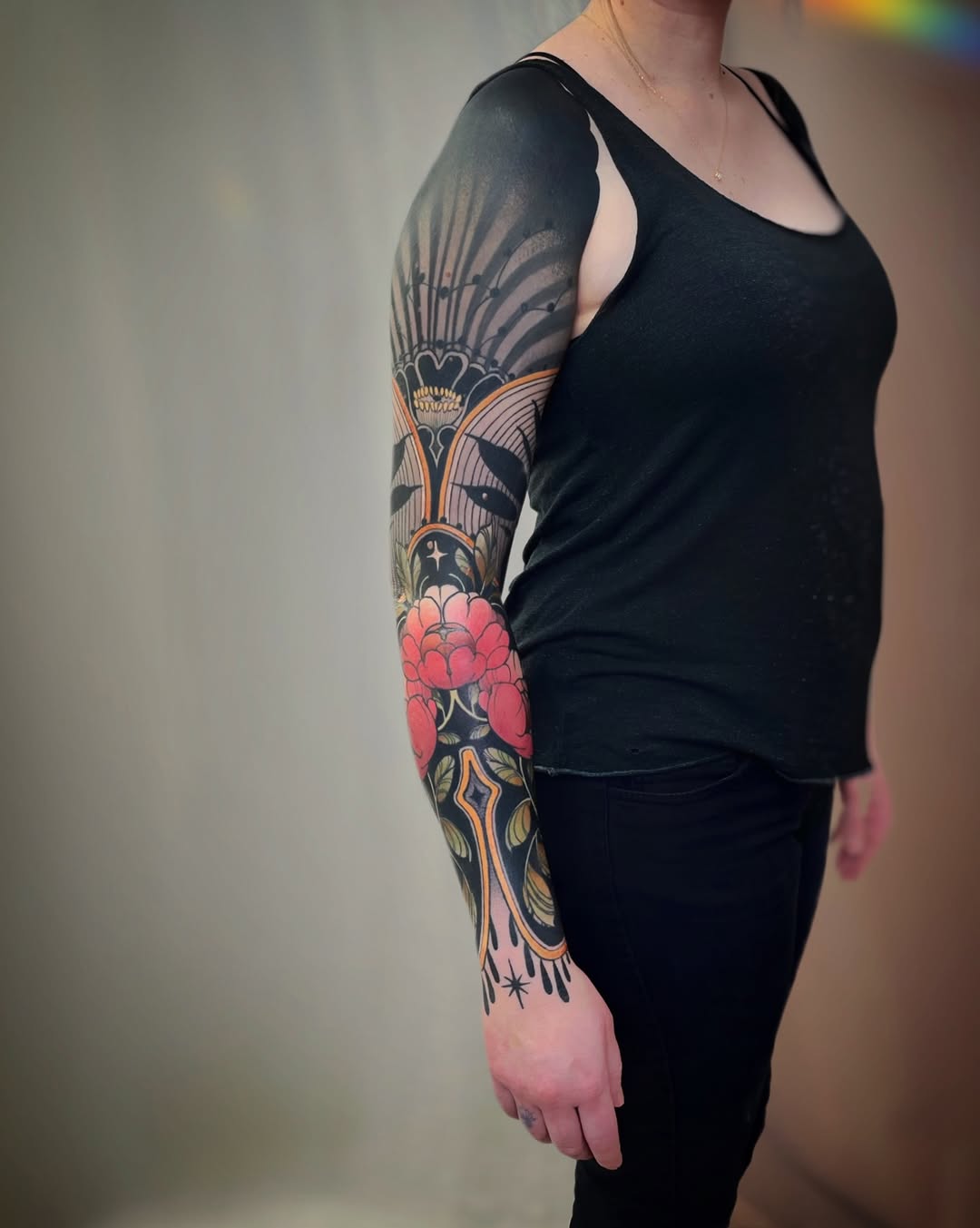 Full right arm sleeve tattoo featuring winged hearts, red flowers, crosses, and ornamental designs in black, red, and gold on a person wearing a black tank top.