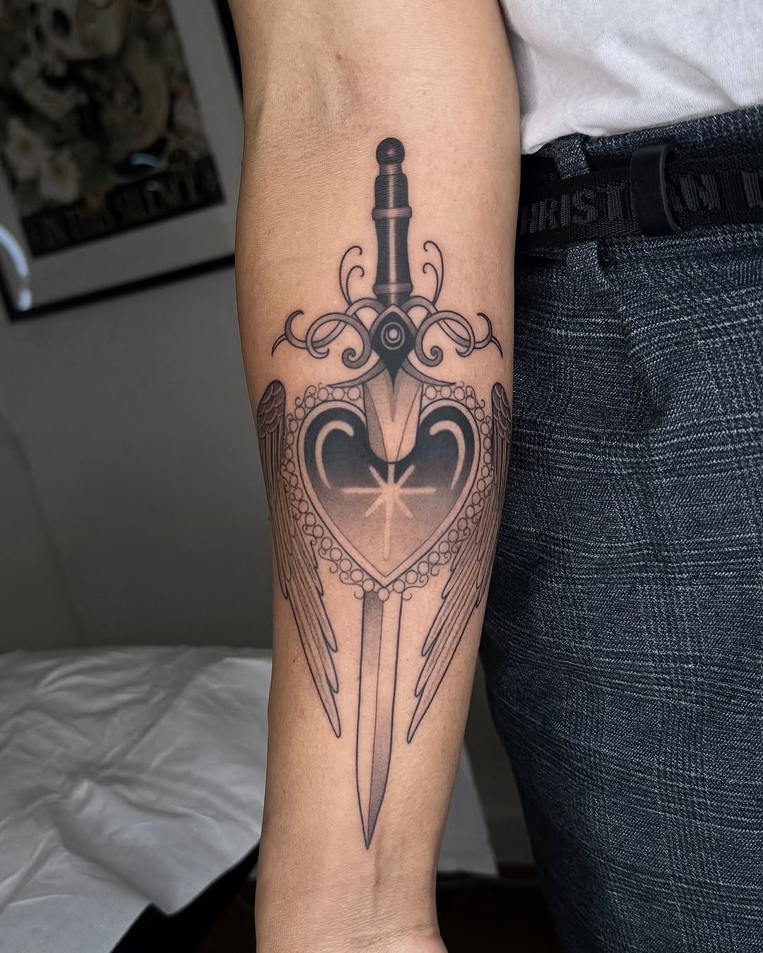 Ornate winged heart with sword and decorative flourishes in black ink on a client's forearm.