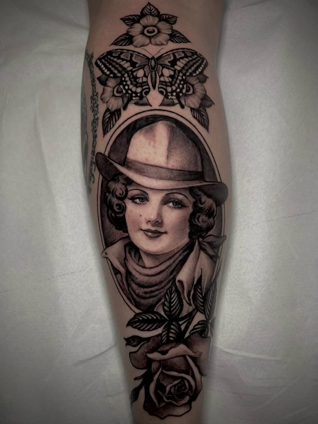 Vertical leg tattoo featuring a portrait of a woman in a fedora hat surrounded by a butterfly, flowers, and rose, rendered in black ink.