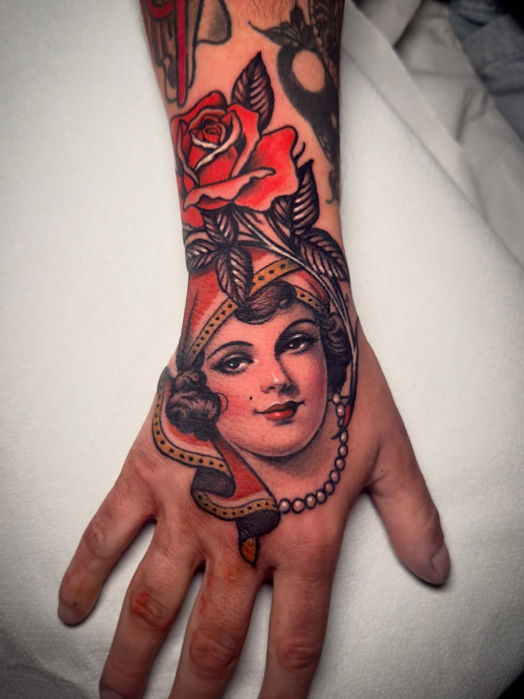 Portrait tattoo of a woman's face with red rose and decorative elements on hand and forearm in red, black, and gold ink.