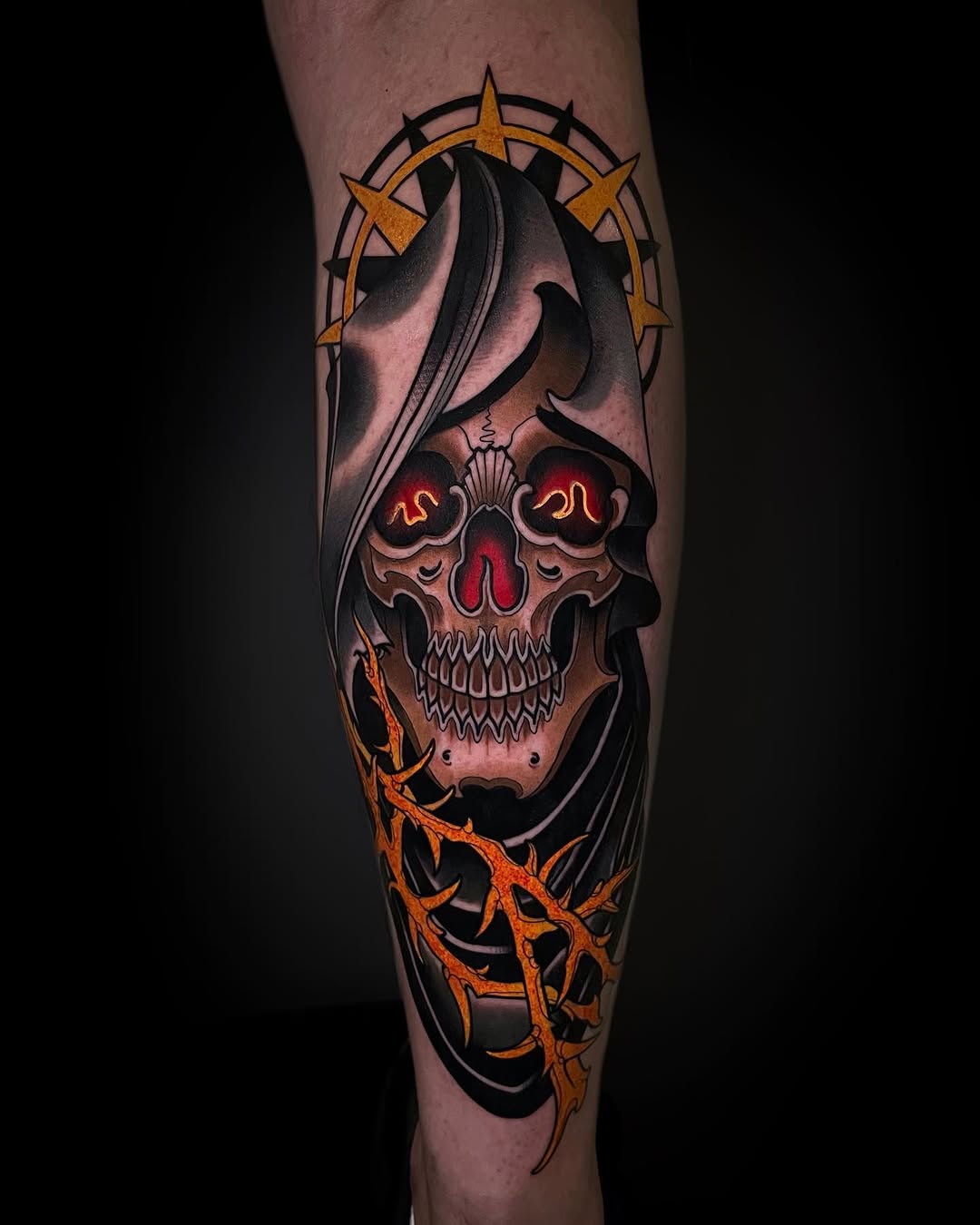 Skull tattoo with glowing red eyes, golden compass star above, black hooded reaper figure, and orange thorned branches below on a leg.