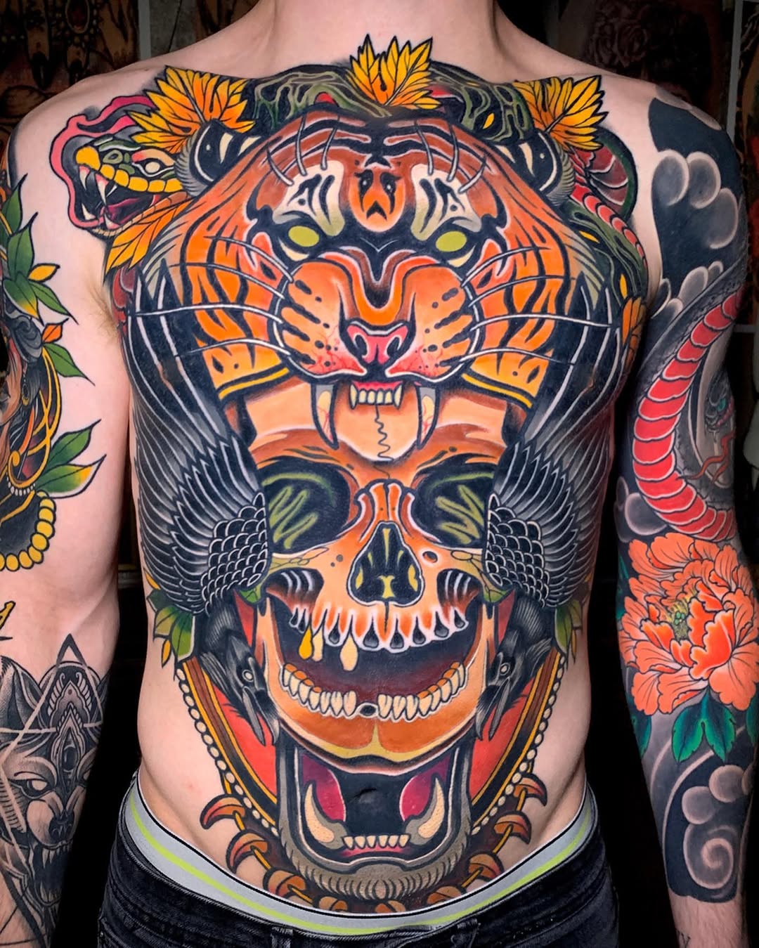 Large chest and torso tattoo featuring a tiger head above a skull with wings, surrounded by flowers and decorative elements in orange, yellow, black, and red.