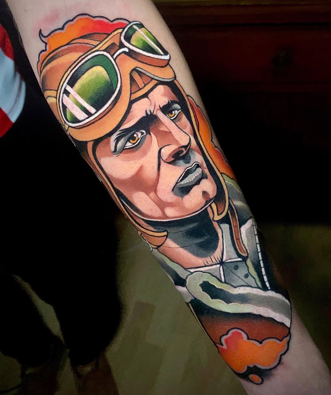 Colorful portrait tattoo of a person wearing orange aviator goggles and flight gear on the upper arm, featuring vibrant orange, green, and skin tones.