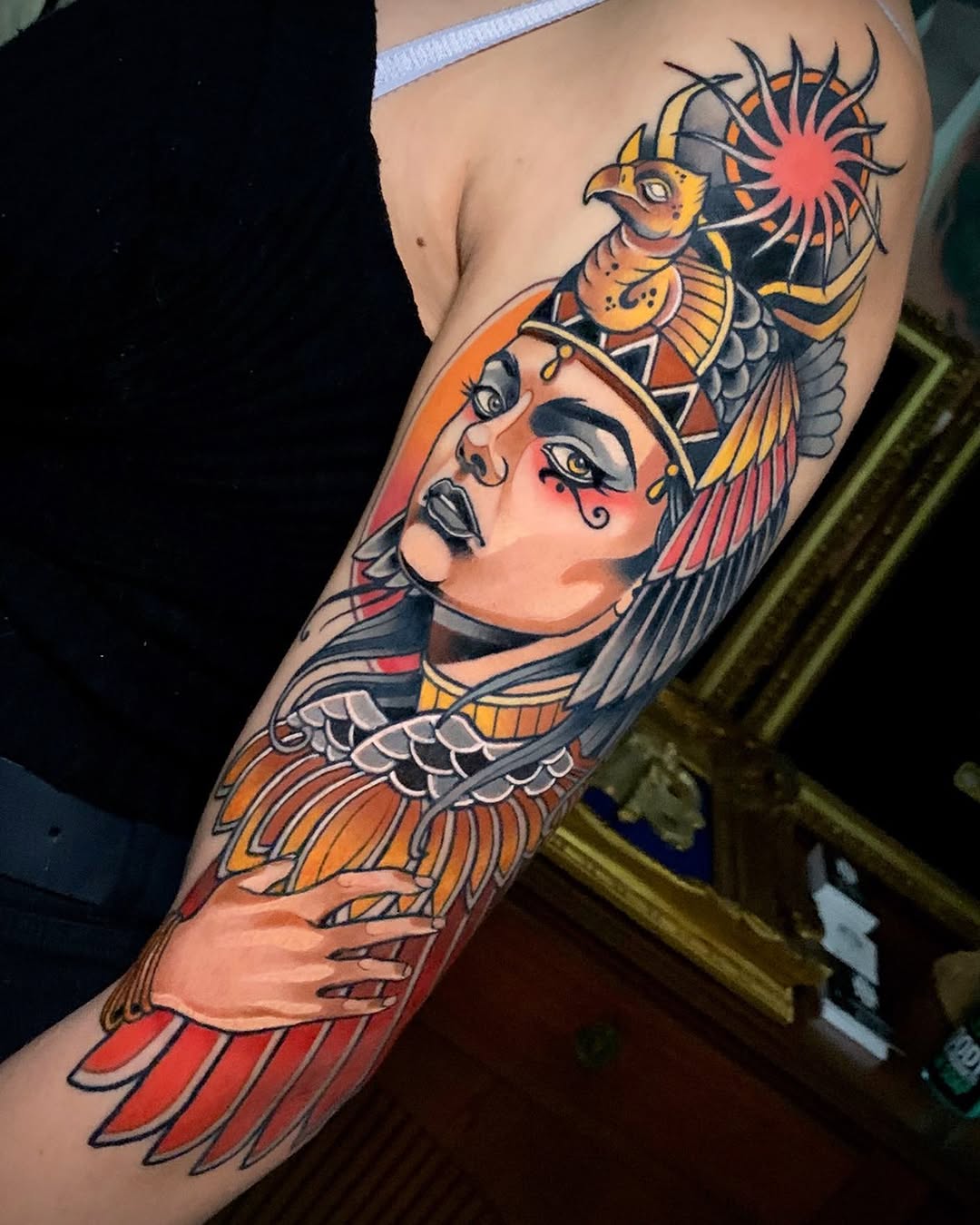 Colorful shoulder and arm tattoo featuring an Egyptian-inspired woman's face with winged headdress, ornate bird, and sun symbol in orange, red, yellow, and black.