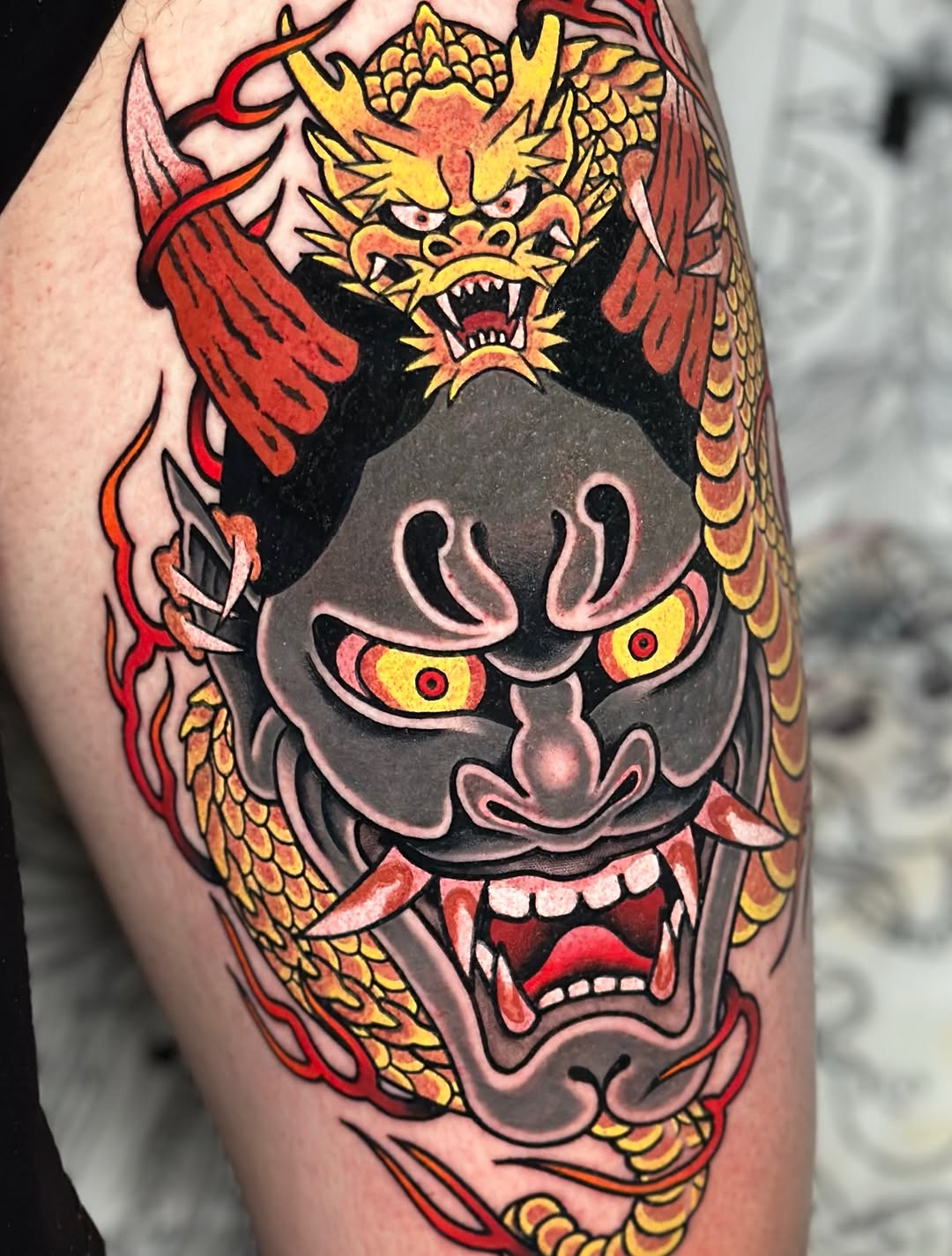 Colorful demon mask and dragon tattoo on thigh featuring yellow, red, and grey tones with detailed facial expressions and scaled wings.